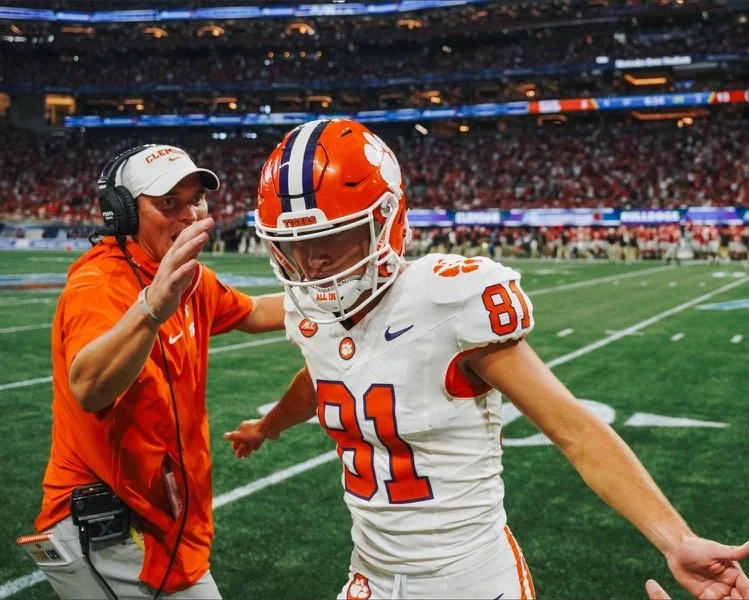 A football player wearing a white and orange uniform with the number 81, practicing on a football field while a coach or staff member in an orange shirt and headset, appears to be giving instructions or encouragement.