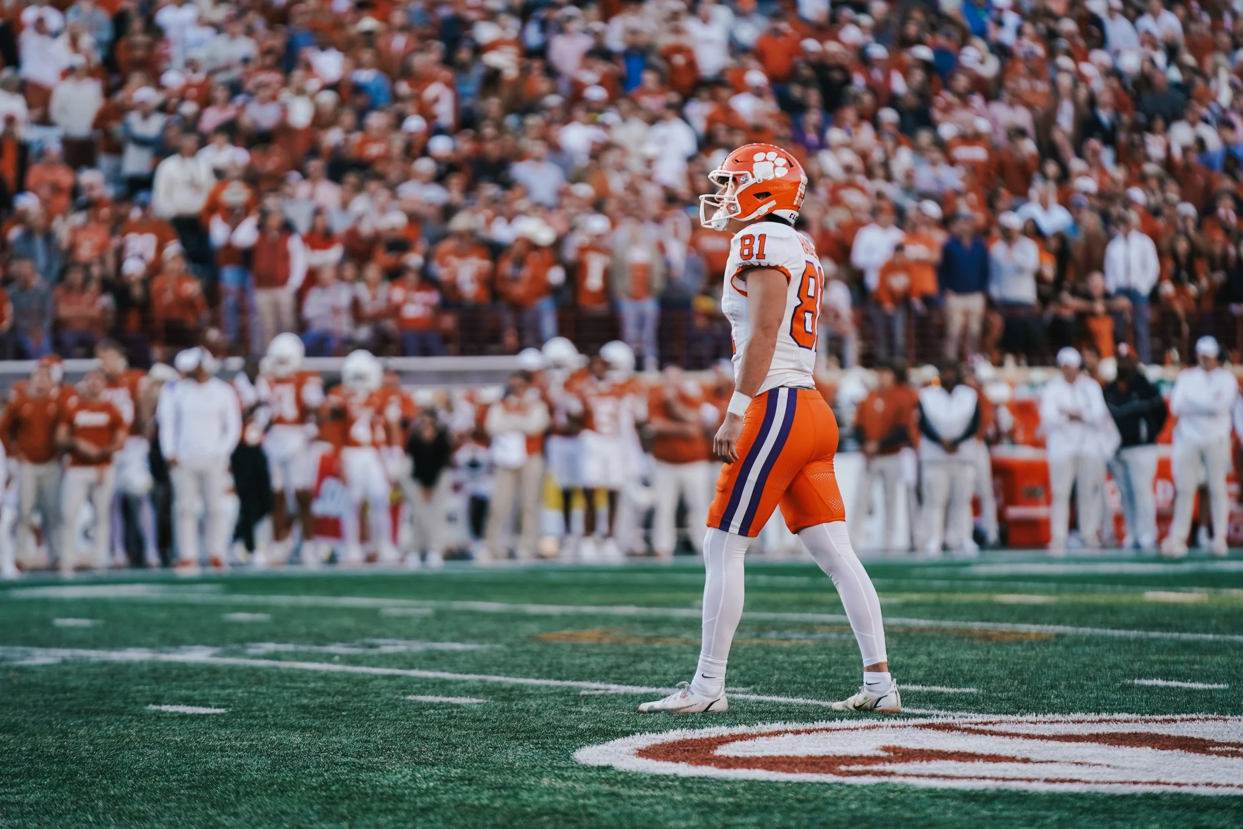 An American football player in orange and white uniform standing alone on the field with a crowd in the background.
