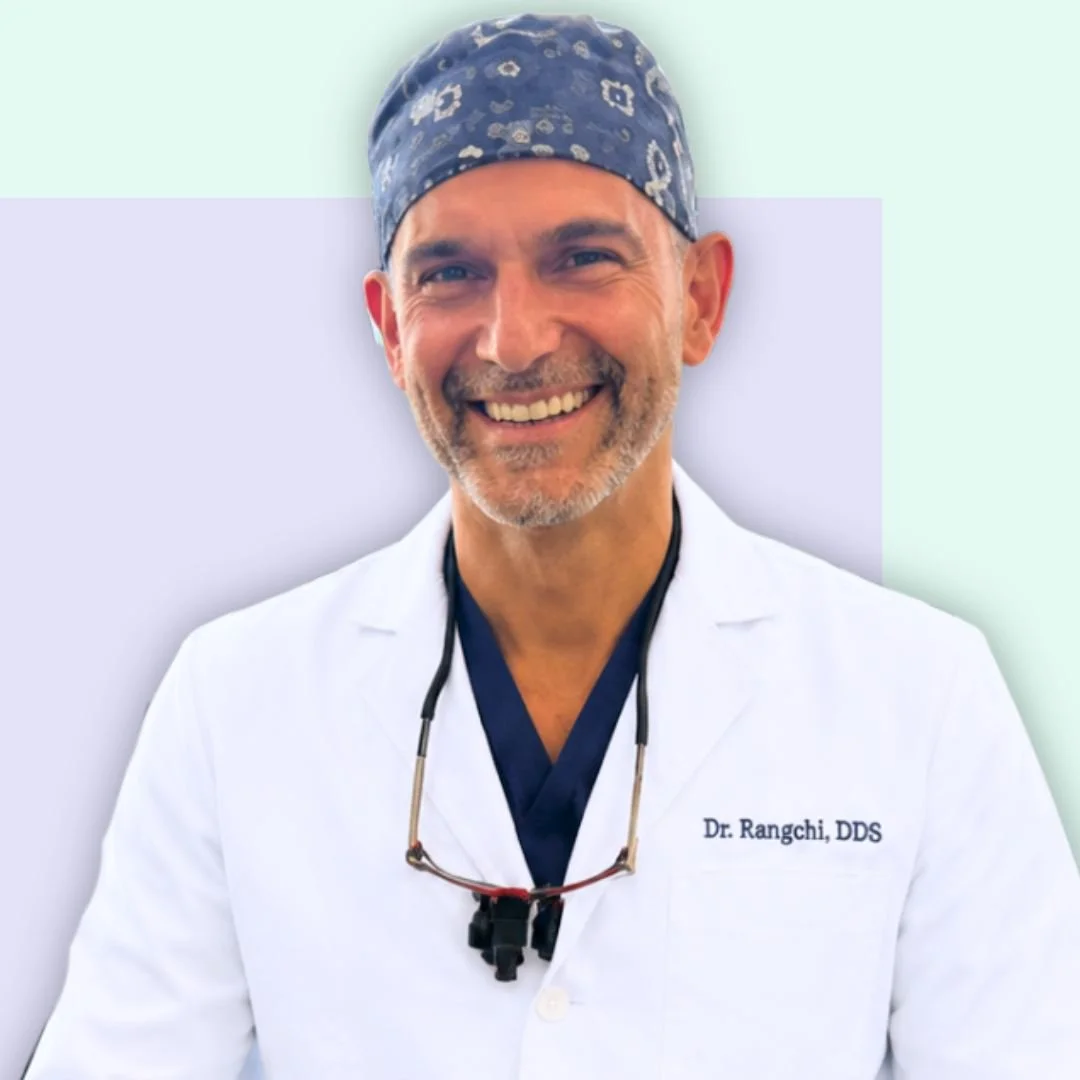 Smiling male doctor wearing scrubs, a surgical cap, and a stethoscope, standing in front of a light-colored background.