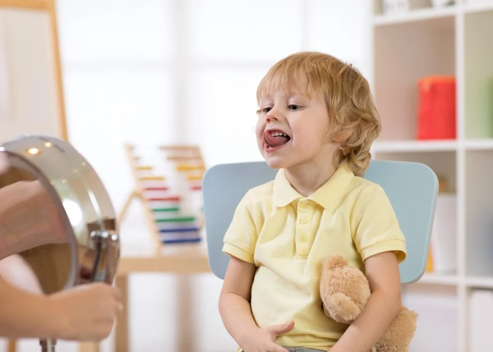 A young boy with red hair sits in a high chair, sticking out his tongue, as a person holds a shiny metal bowl in front of him in a bright room.