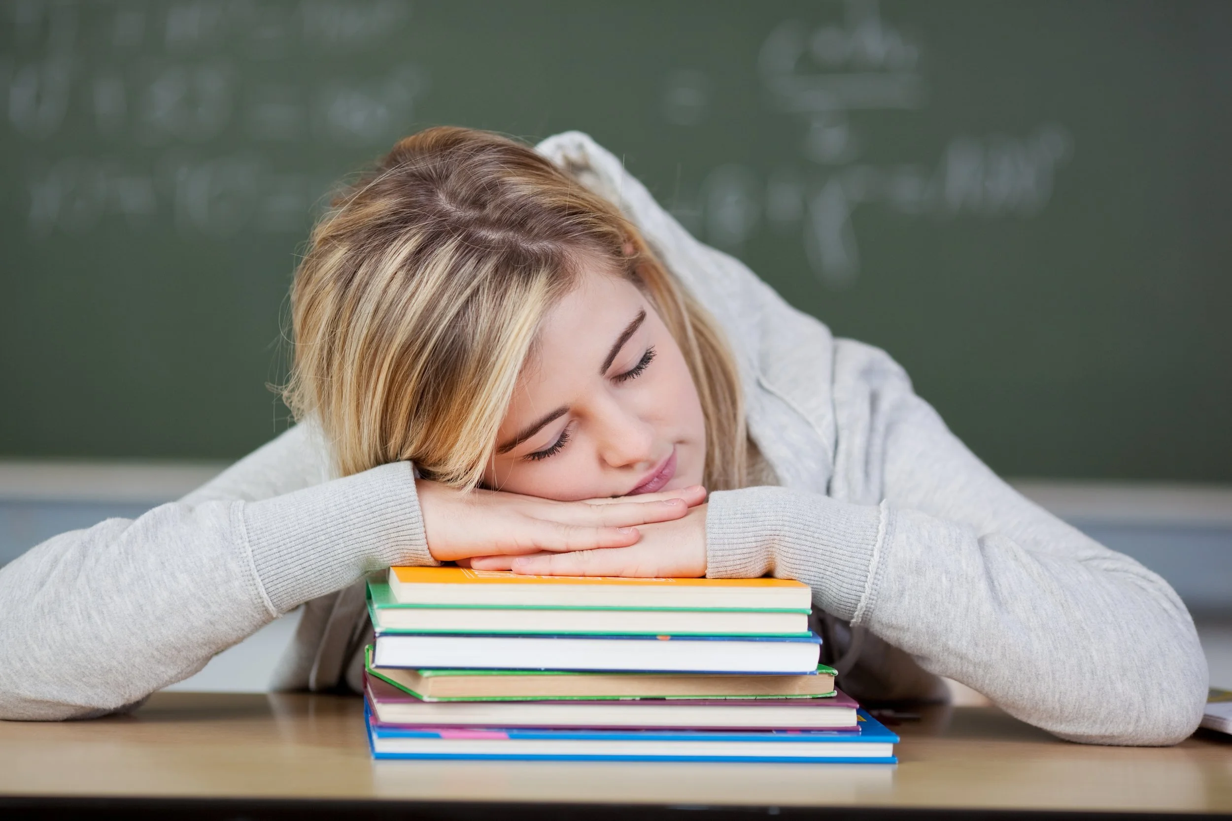 A young woman with blonde hair resting her head on a stack of colorful books on a desk in a classroom.