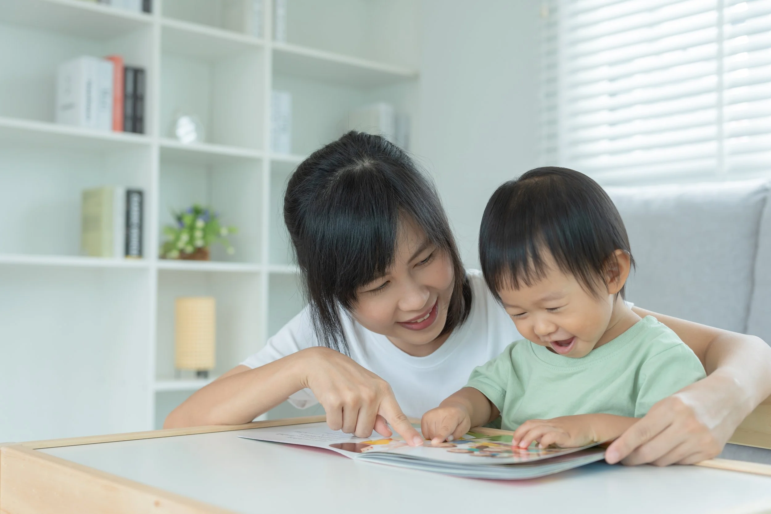 A woman and a young child are looking at a colorful book together at a table in a bright room with white shelves and blinds