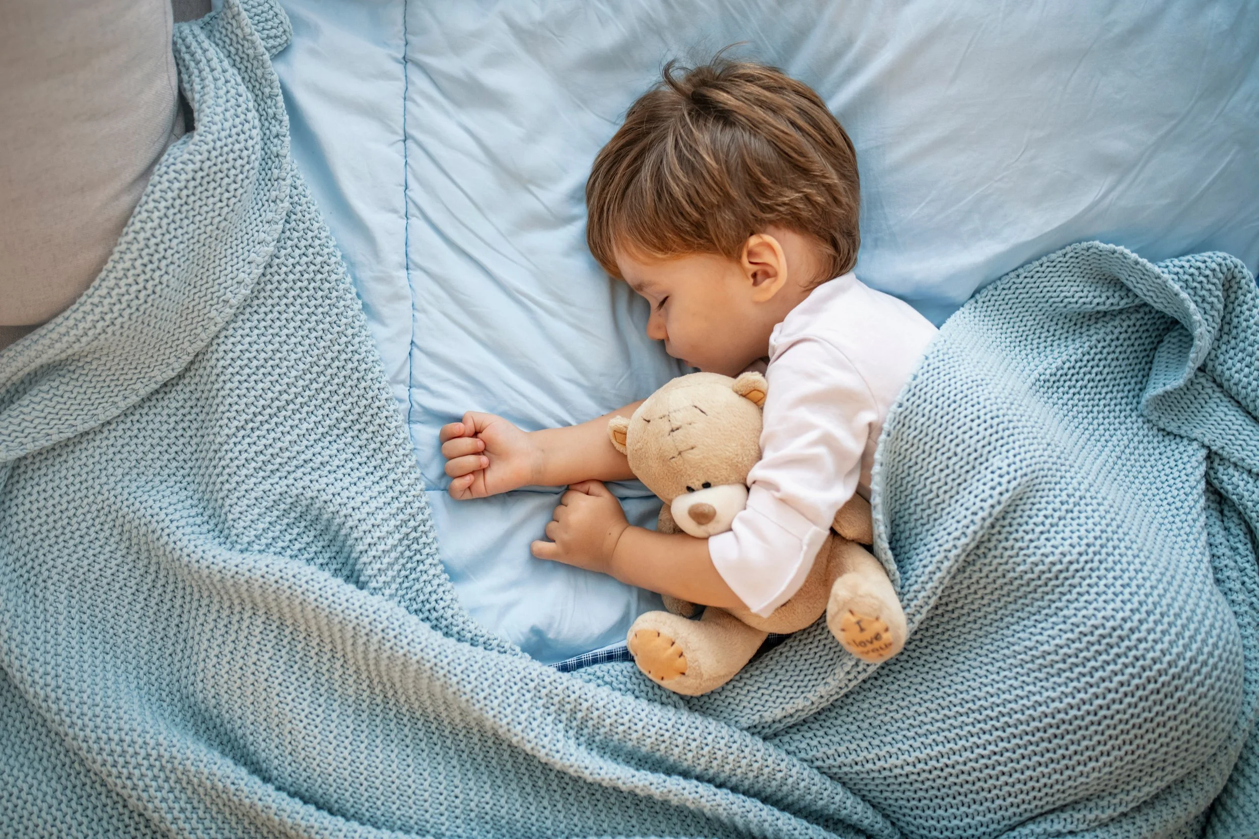 Young boy sleeping in bed, hugging a teddy bear, covered with blue knitted blanket, resting on a light blue pillow.