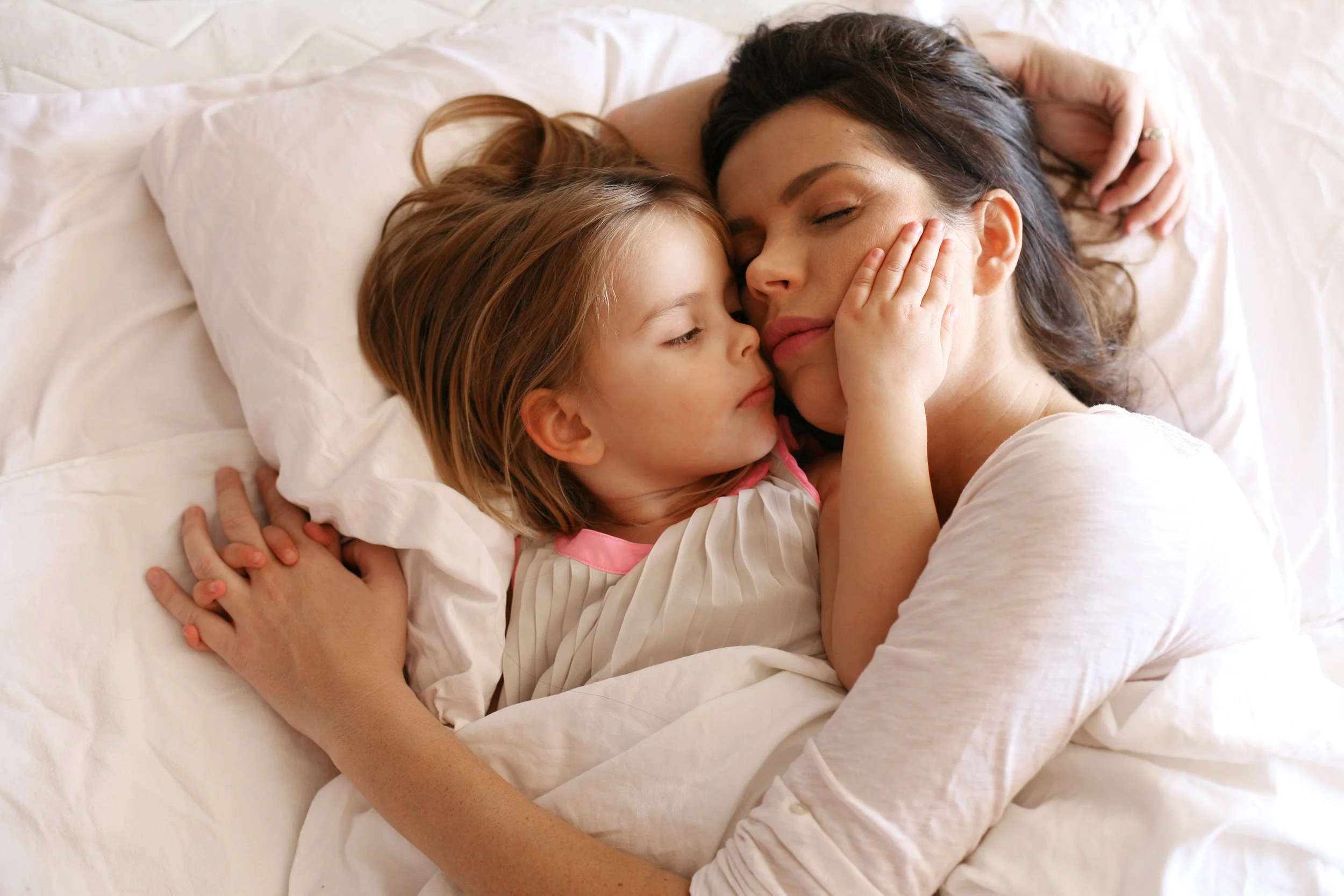 A woman and a young girl lying on a bed, touching faces and eyes closed, sharing an affectionate moment.