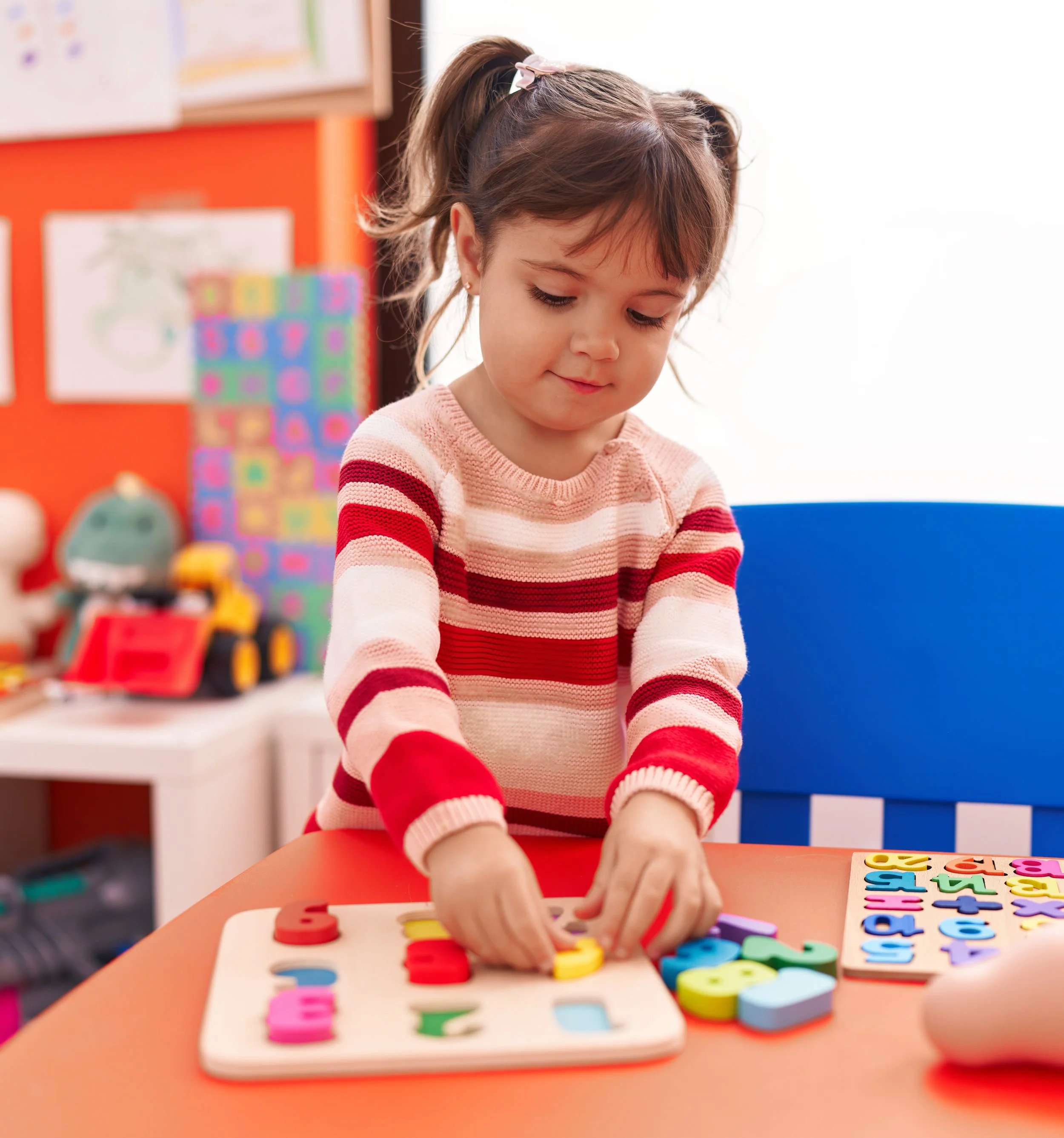 Young girl playing with colorful foam alphabet letters at a red table in a classroom.