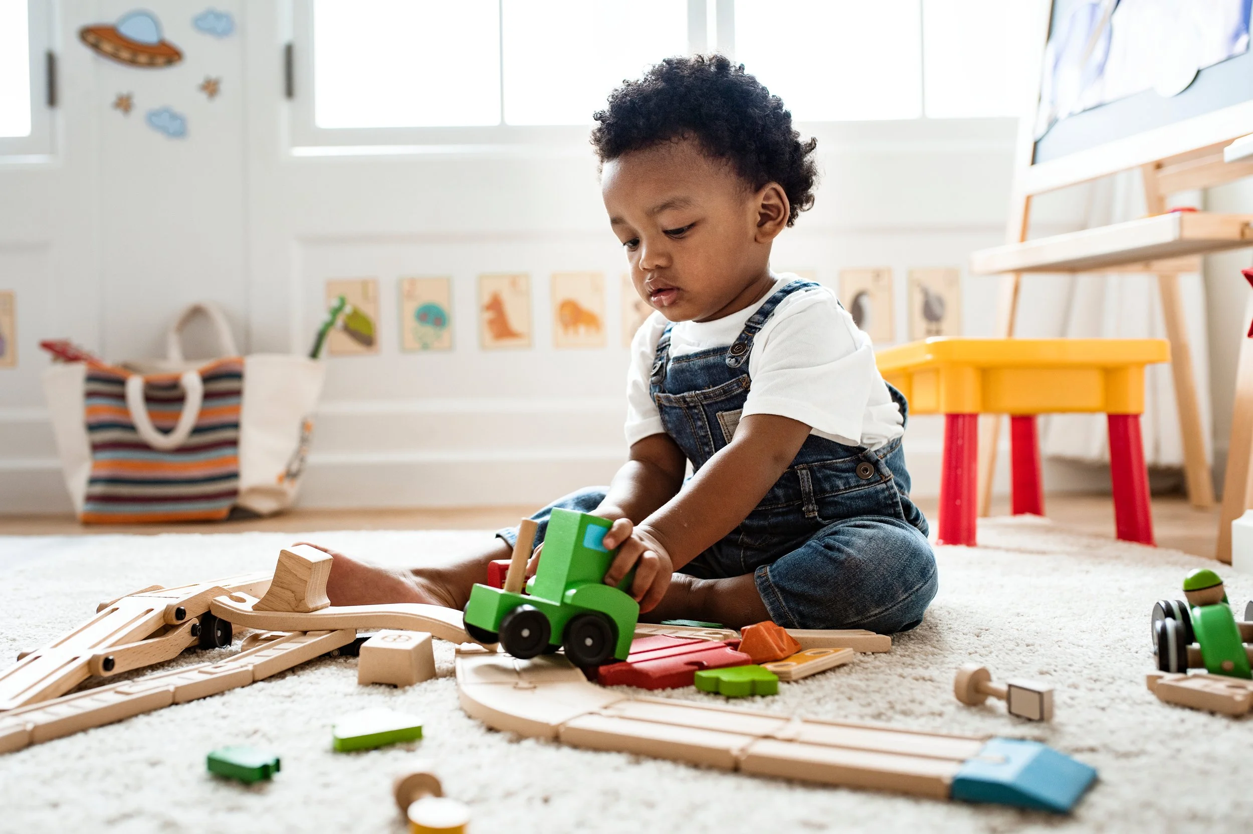 A young child playing with wooden train set on a beige carpet in a bright, decorated playroom.