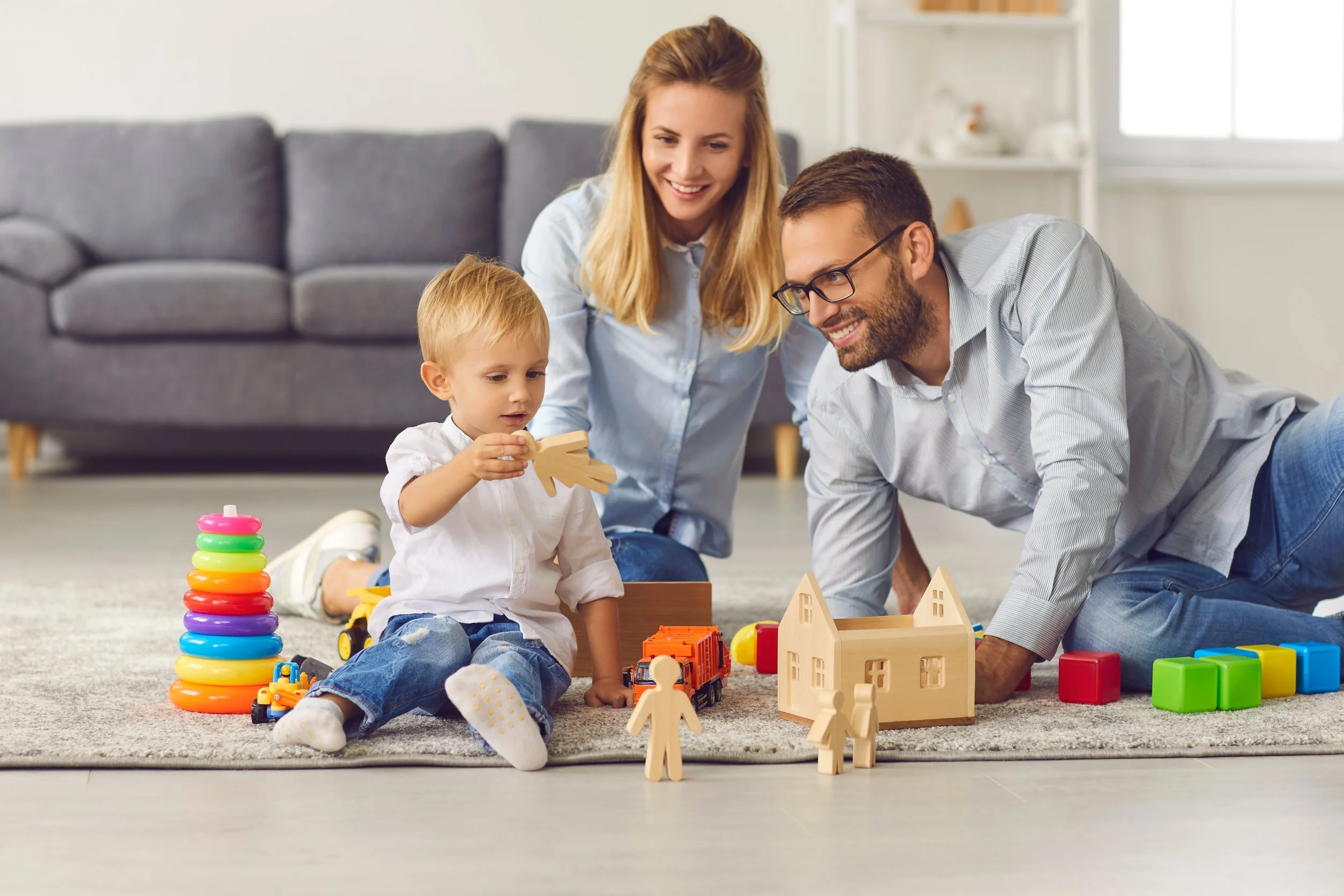 A family playing with toys on the living room floor, including a stack of colored rings, wooden figurines, and a toy house, with a gray sofa and shelves in the background.