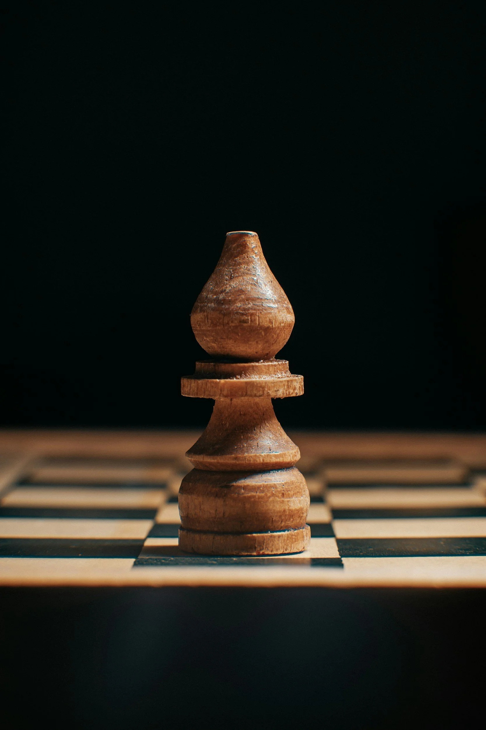 A close-up of a wooden chess piece, a pawn, placed on a chessboard against a dark background.
