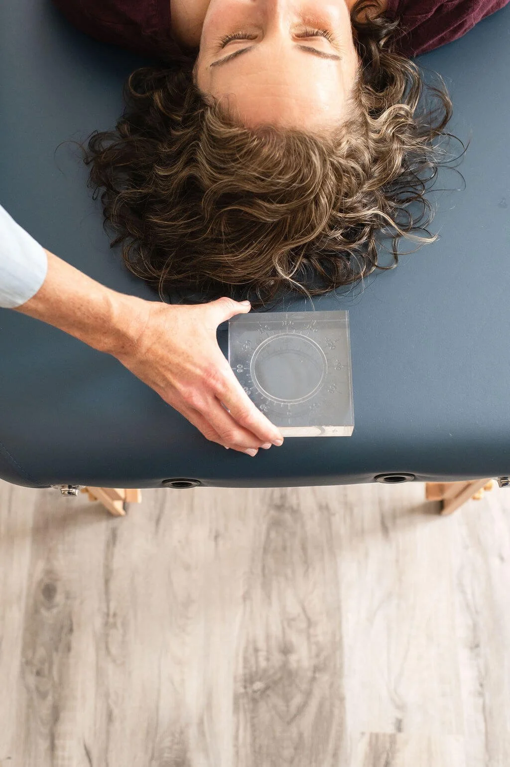 A client with curly brown hair lays face up on a blue treatment table, while Caitlin places a transparent circular measurement tool near their head during an autonomic response testing visit.