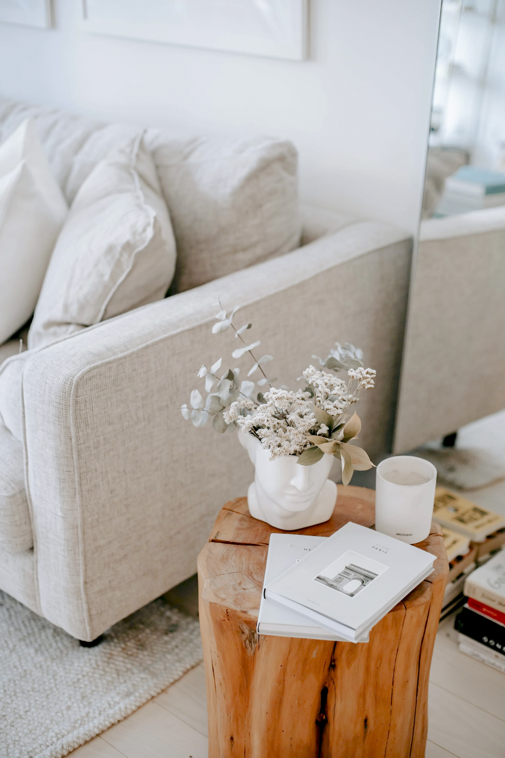 Living room with beige sofa, white pillows, a small wooden side table with a floral arrangement in a white face-shaped vase, hardcover books, and a candle.