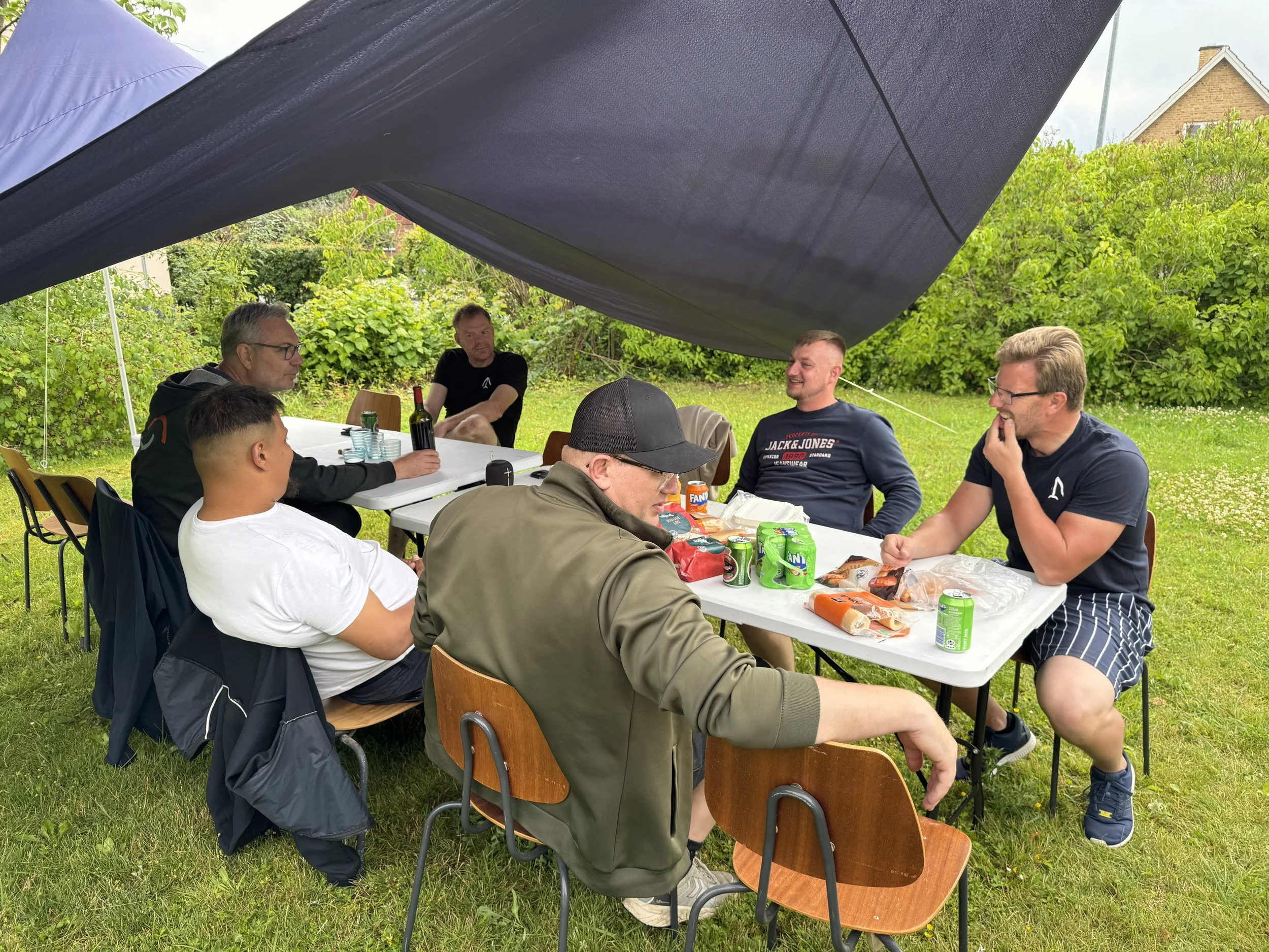 Gruppe af seks mænd sidder rundt om et bord under et stort skyggenet, nyder en grillmiddag med sodavand og snacks i en baghave.