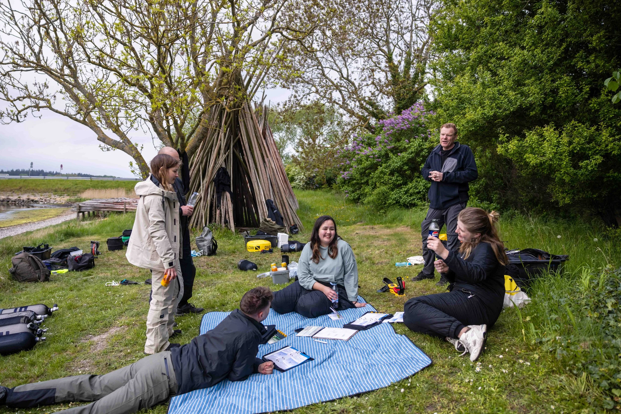En gruppe mennesker sidder og står på en græsset område med buske og træer, med en lejrstillingsstruktur lavet af grene i baggrunden. De har campingudstyr og regnjakker, og nogle ser ud til at være i en samtale eller aktivitet.