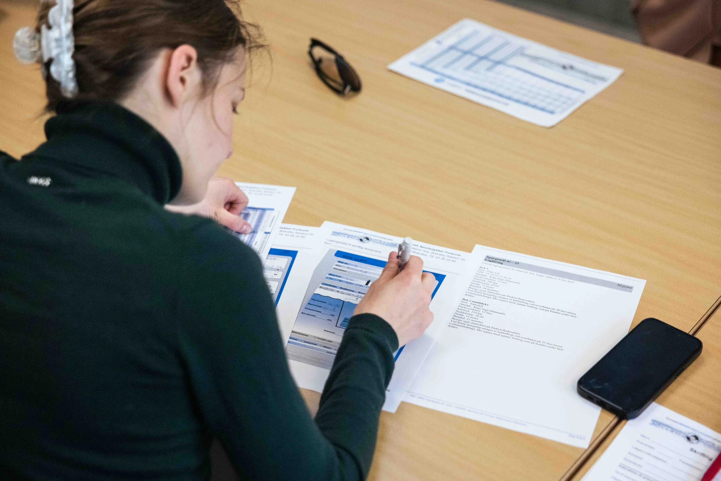 En person sidder ved et bord, skriver på papirer med et blæk. Der er en smartphone og flere papirer på bordet.