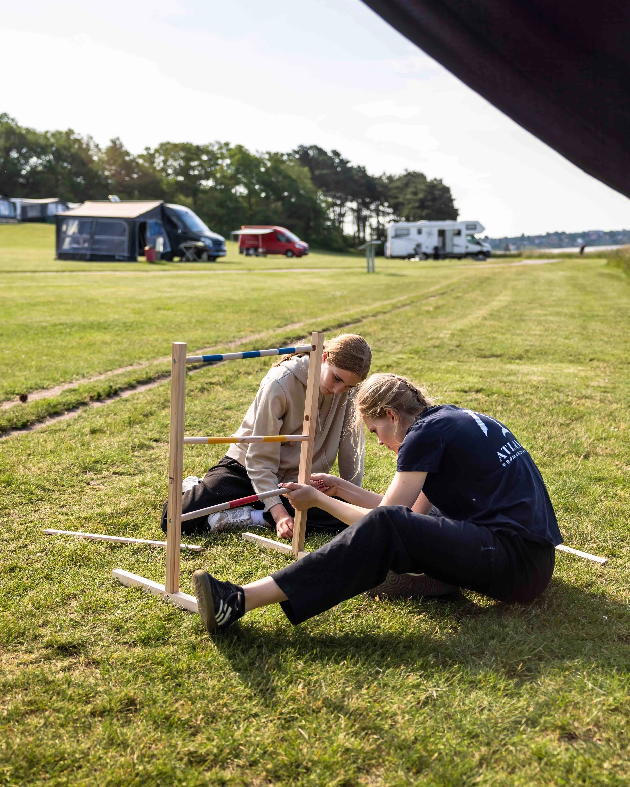 To unge kvinder samler et stykke legeredskab på en græsplæne med campingvogne i baggrunden.