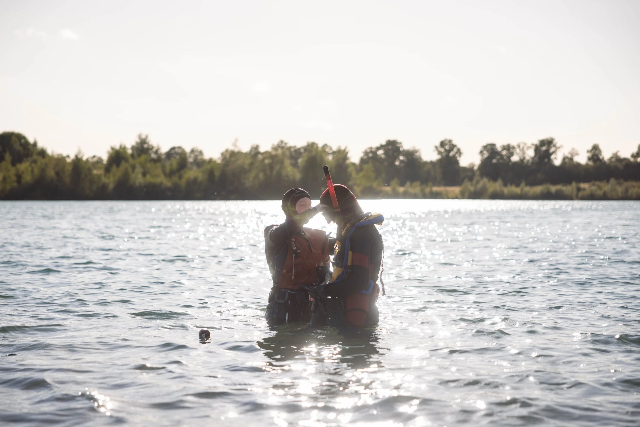 To personer i våddragt og snorkeludstyr i vandet, der hjælper hinanden med at justere udstyr, med sollys reflekterende vand, og en grøn skov i baggrunden.