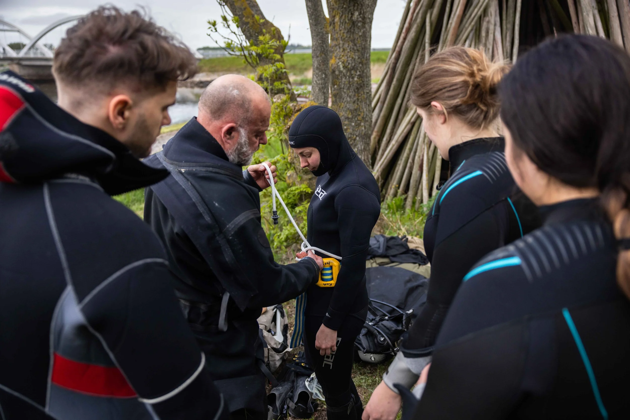 En gruppe dykkere får instruktioner fra en instruktør, der viser dem udstyr og hjælper med forberedelse i udendørs omgivelser.