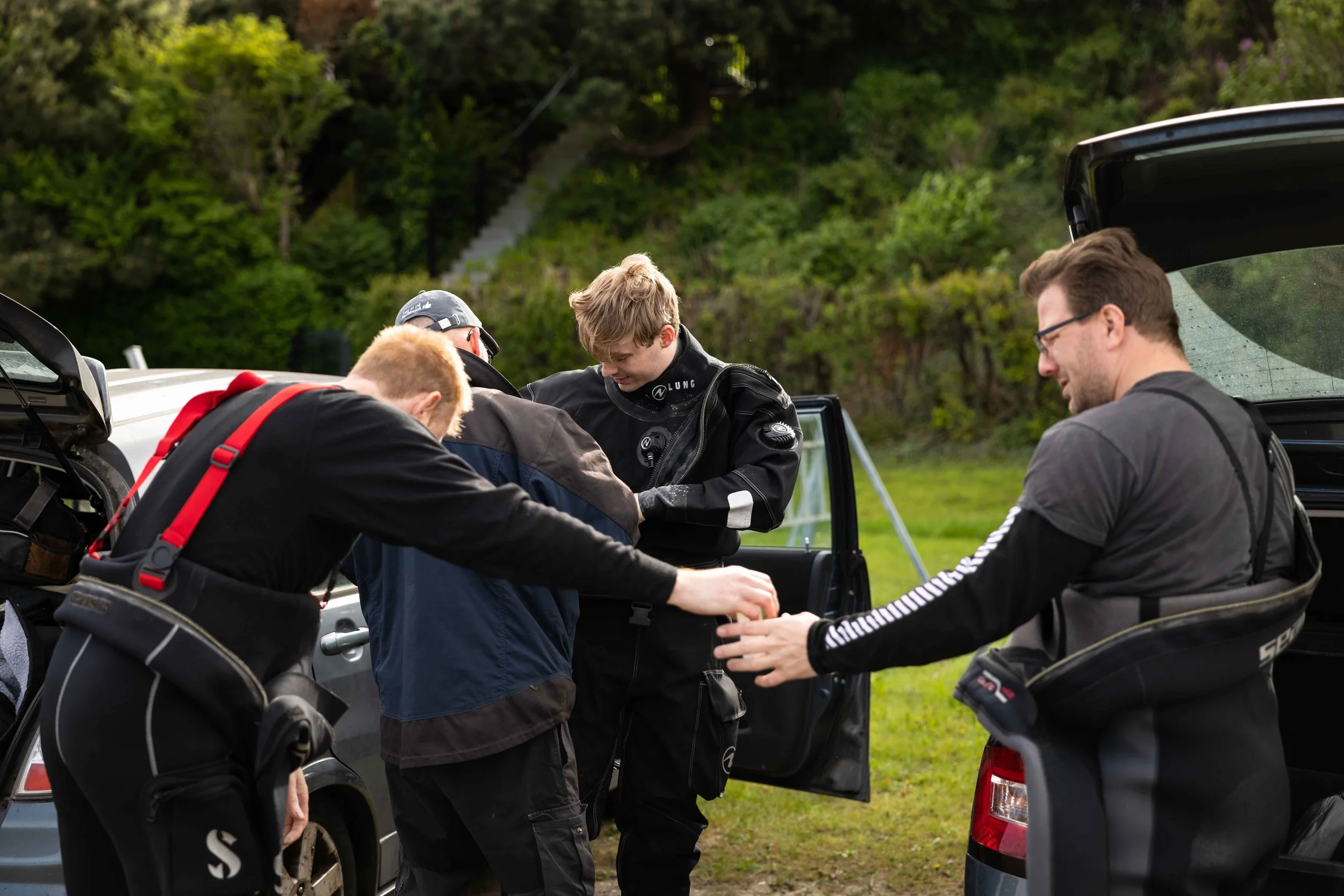 Fire personer, to mænd og to kvinder, forbereder sig på motorcykling ved biler på en græsplæne med grønne træer i baggrunden. De er iført motorcykeltøj og har udstyr.