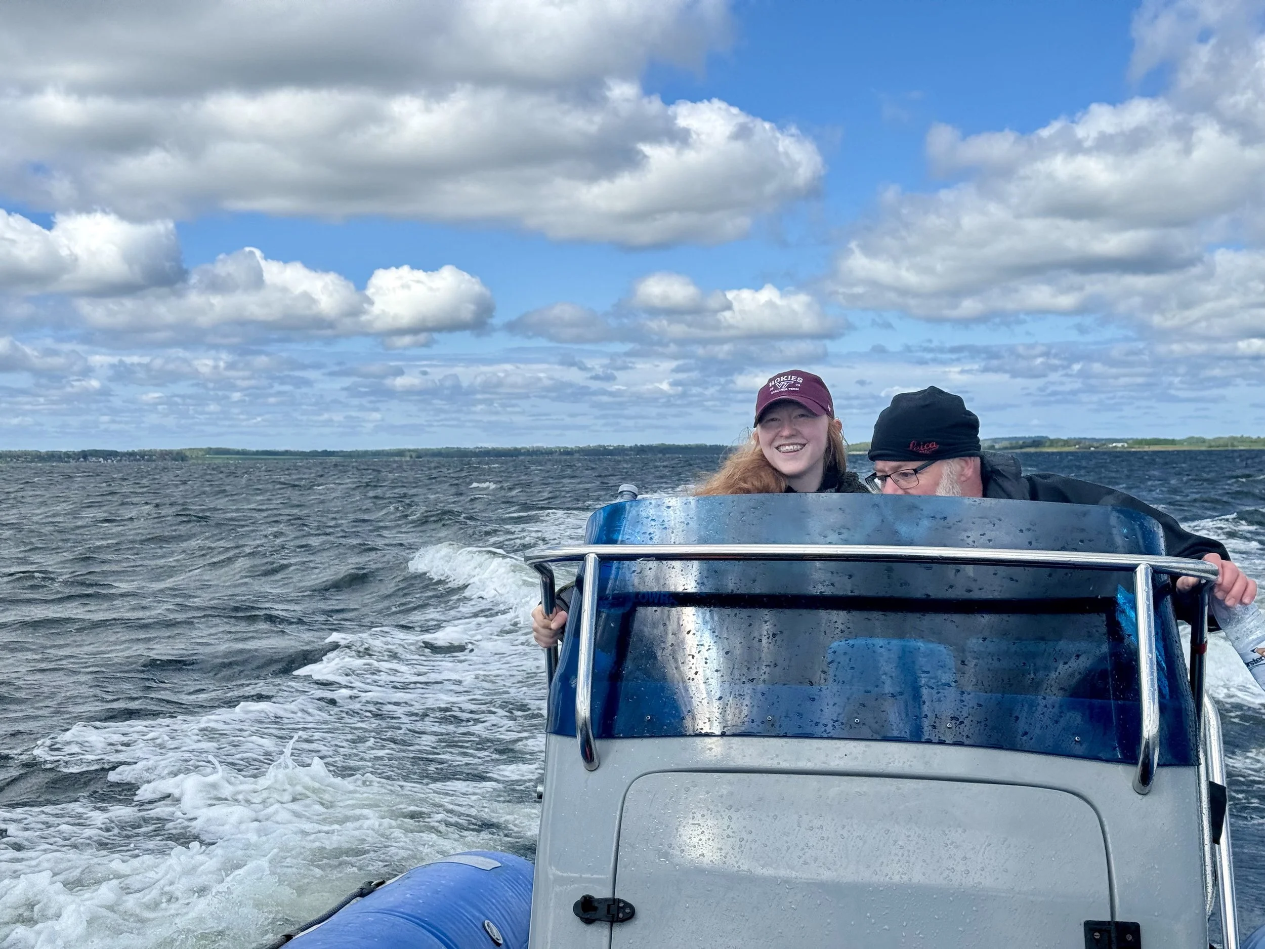 To personer på en båd på vandet, en kvinde med rød hår og en mand med briller, begge smilende. Båden bevæger sig hurtigt på vandet under en skiftende skyet himmel.