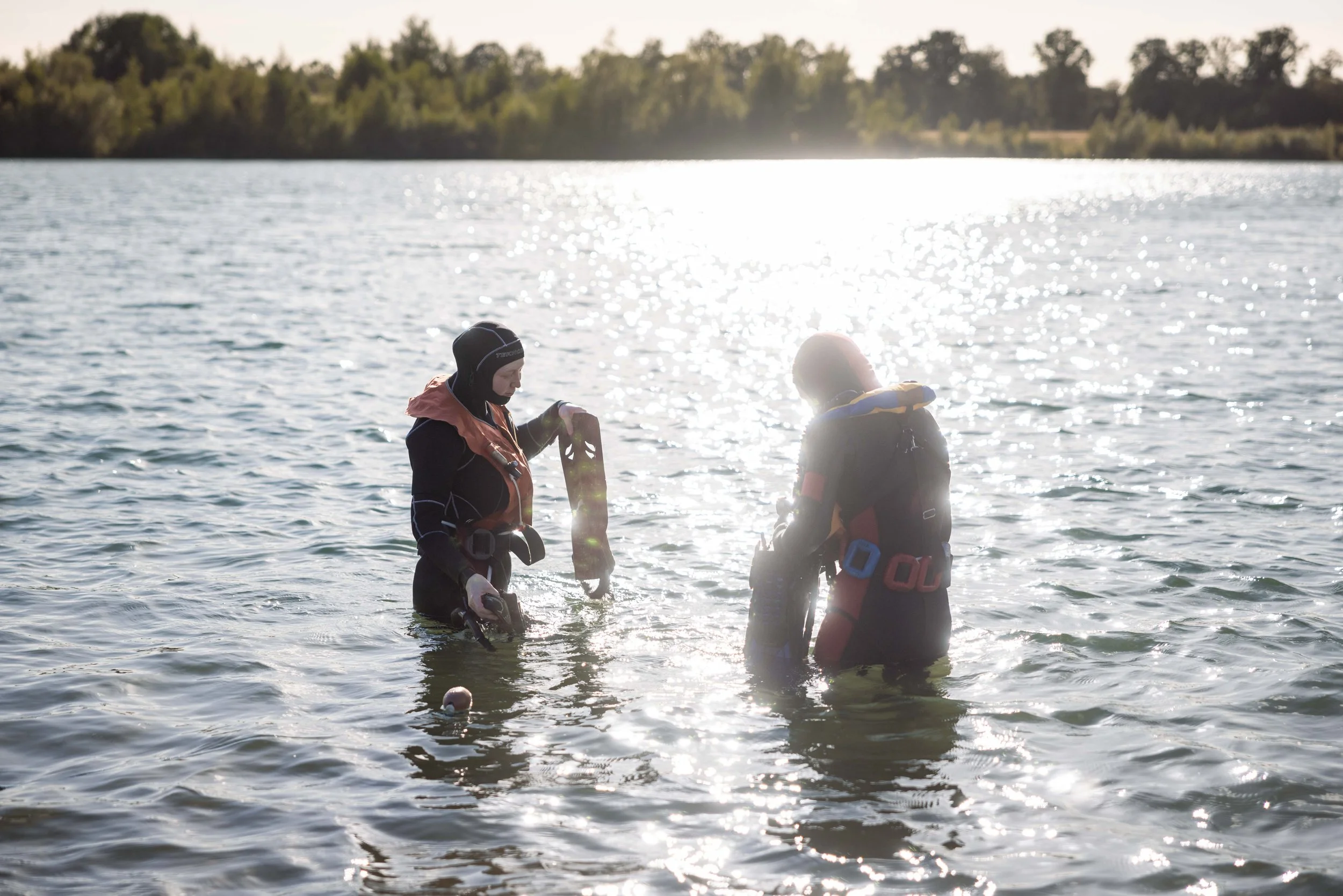 To personer i våddragter står i en sø og ser på noget, de har i hænderne, med solen der skinner på vandoverfladen.