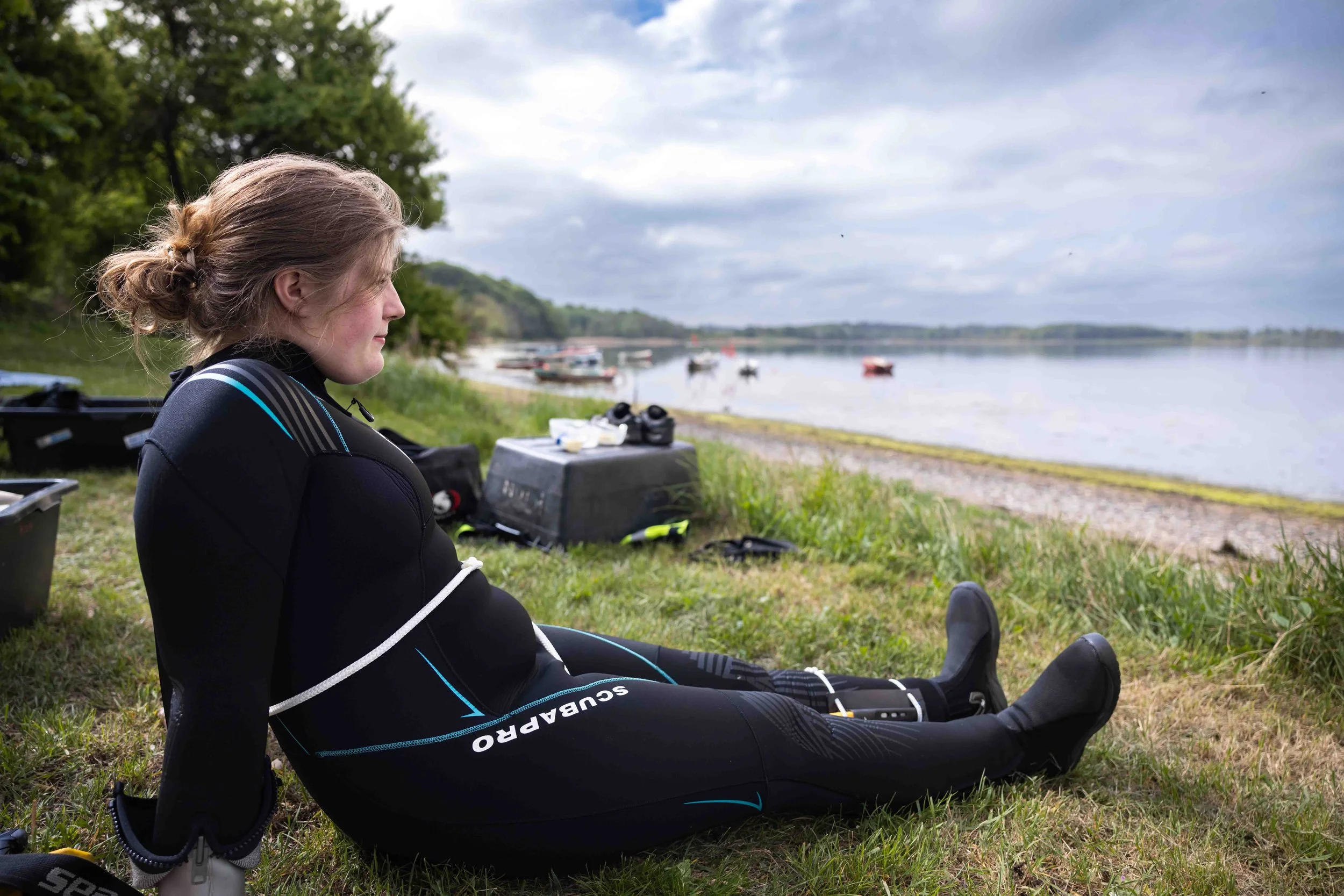 Kvinde i en våddragt sidder på græs ved en sø, omgivet af udstyr, med vand og små både i baggrunden under en overskyet himmel.