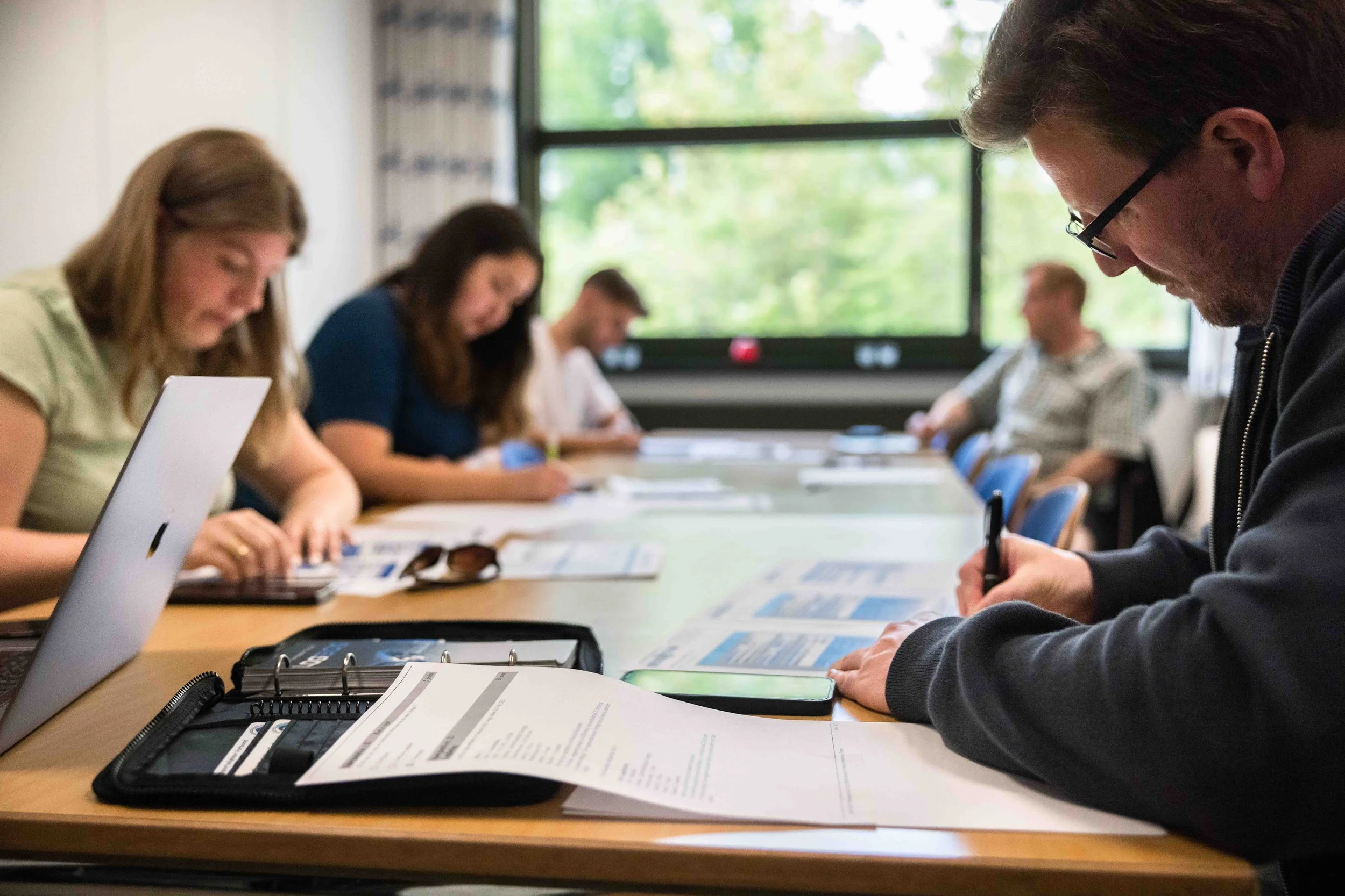 Mennesker sidder ved et langt bord i et mødelokale, læser dokumenter og skriver noter, med store vinduer ud til grønne træer i baggrunden.