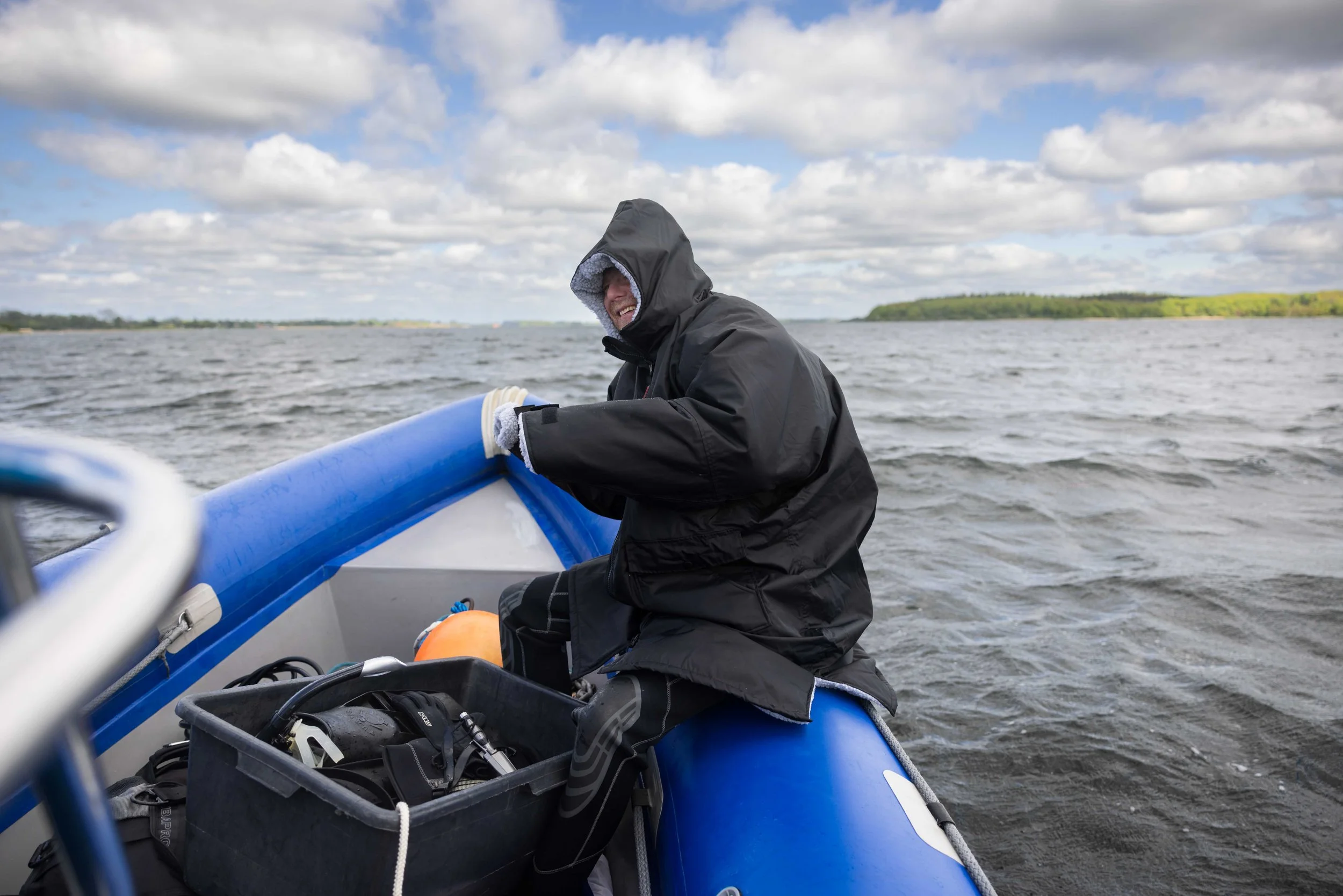 En person i en sort jakke og handsker sidder i en blå oppustelig båd på åbent vand under en skypysslet dag.