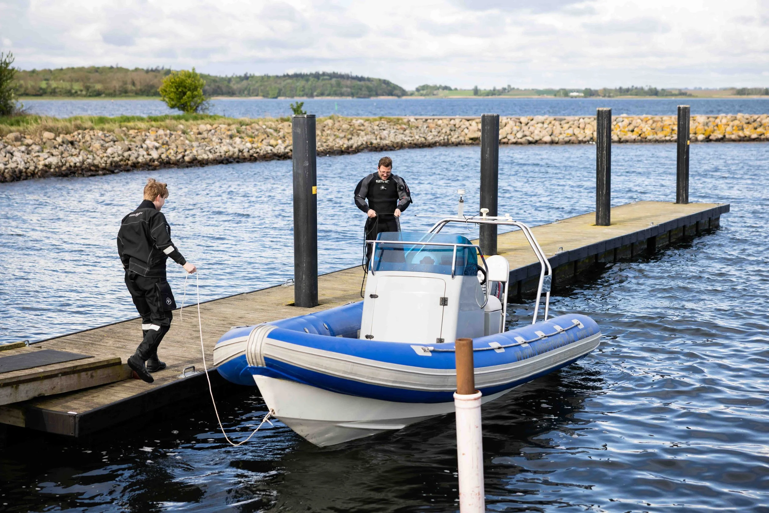 To personer i redningsdragter ved en båd, der bliver slept op på en kaj ved en sø, med en stenmur og grønne træer i baggrunden.