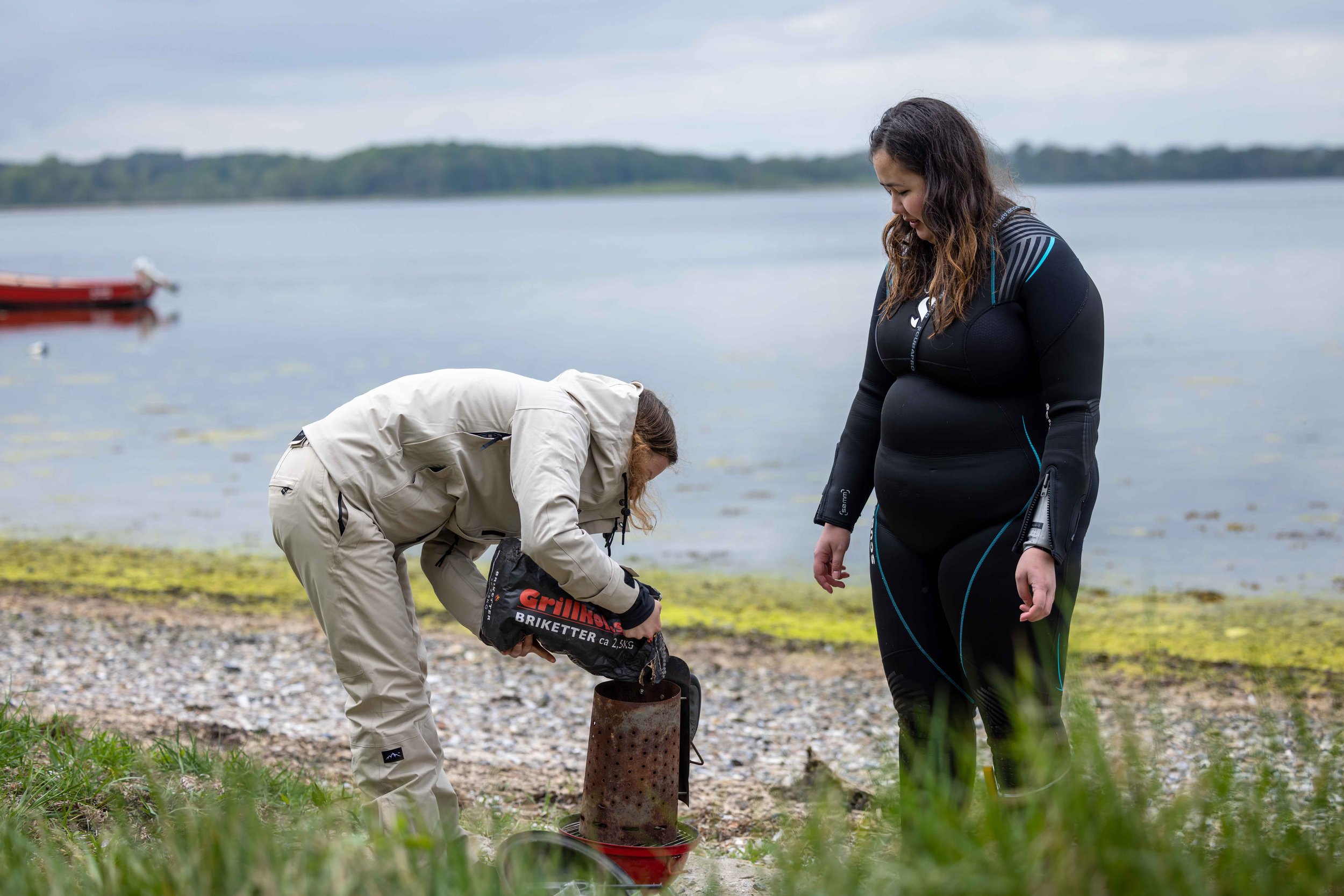 To kvinder ved søen, en iført divers udstyr og en anden i jakke, forbereder en fyr til at affyre en raket til at sprænge en stålboks, en såkaldt bål, på stranden.