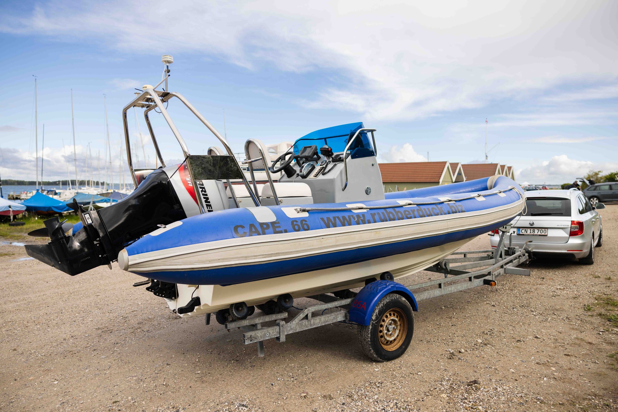 En blå og hvid redningsbåd på trailere med motorkraft, parkeret på en grusjord ved en havn med både og småhytter i baggrunden.