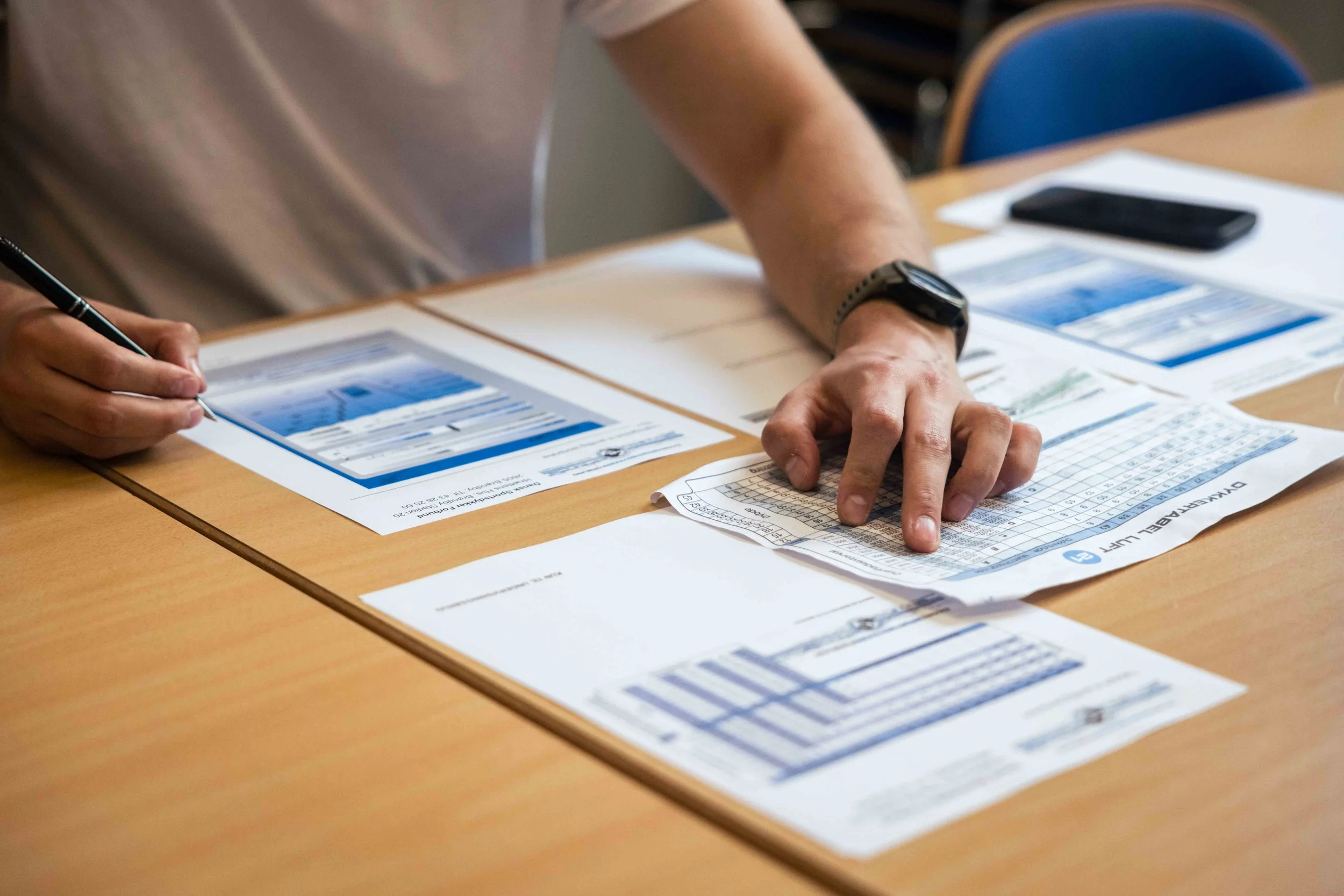 Person, der udfylder et skattepapir på et bord, med en mobiltelefon og andre dokumenter.