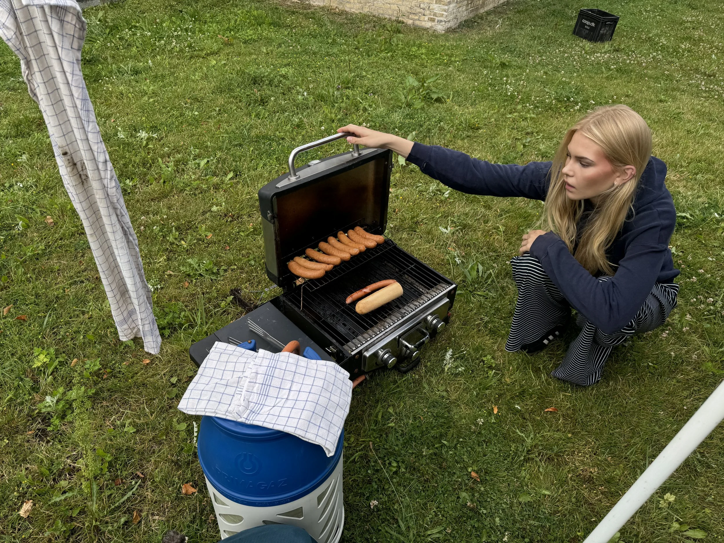 En ung kvinde med blond hår, iført en mørk jakke og stive bukser, kigger på grill, hvor der er pølser og en hotdog. Hun er udendørs på en grøn græsplæne med en hytte eller mur i baggrunden.