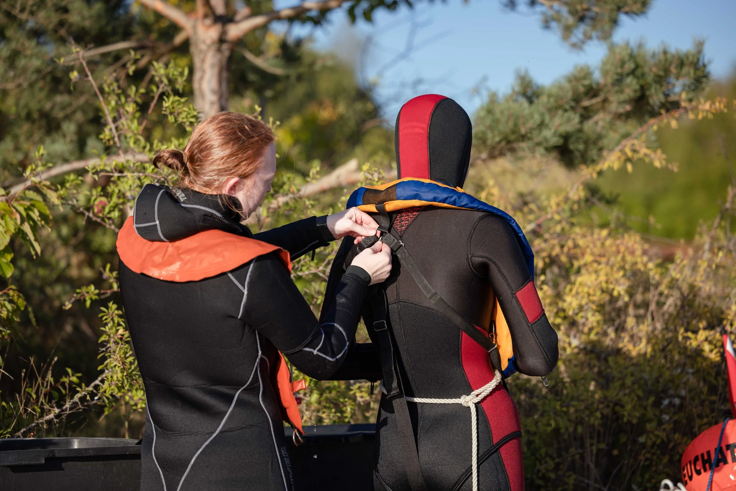 En kvinde, der hjælper en person klædt i en varm rød og sort dragt med at tage noget, måske en rygsæk, på en udendørs beliggenhed med træer og buske i baggrunden.