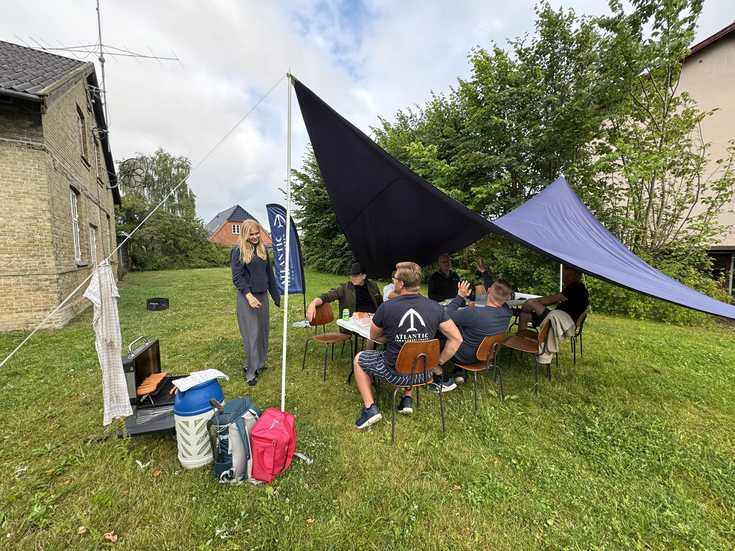 Gruppe mennesker sidder og står under en stor tarp i en have, hvilket ser ud til at være en sammenkomst eller picnic, med grilludstyr og mad.