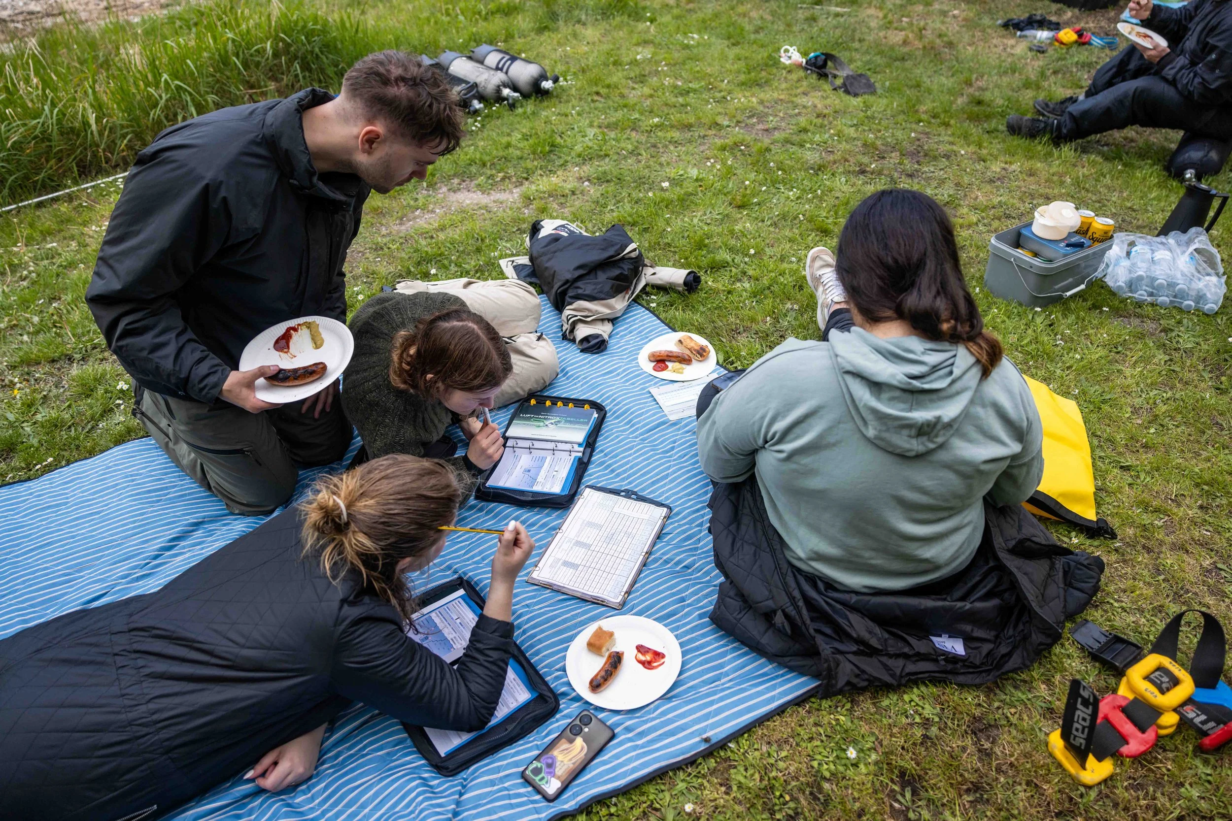 Gruppemedlemmer sidder og står på et tæppe i naturen med mad, udstyr og tabletter, mens de arbejder eller hygger sig.