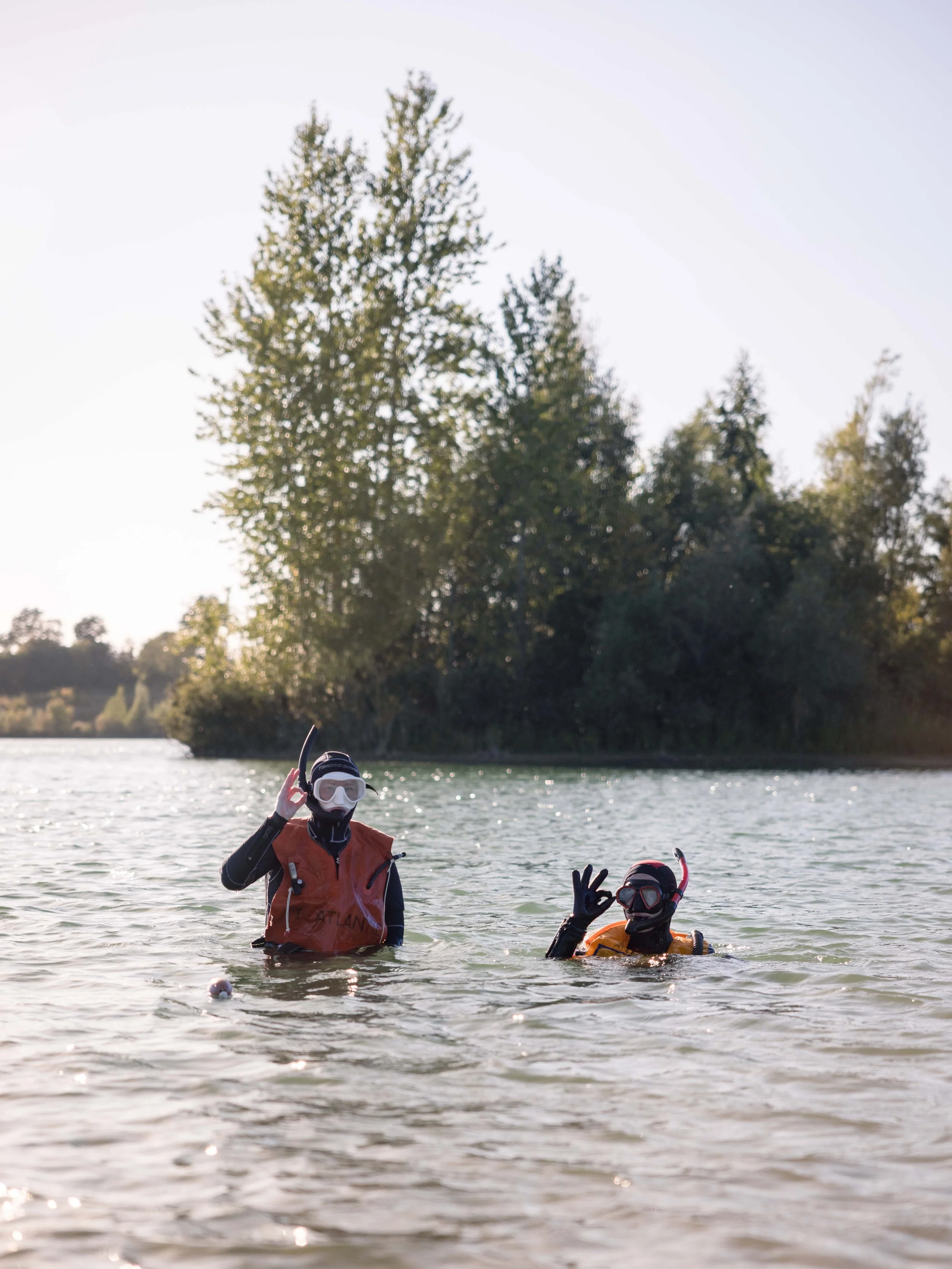 To personer i dykkermaske og tørdragt står i en sø eller flod og giver OK-tegn, med træer i baggrunden.