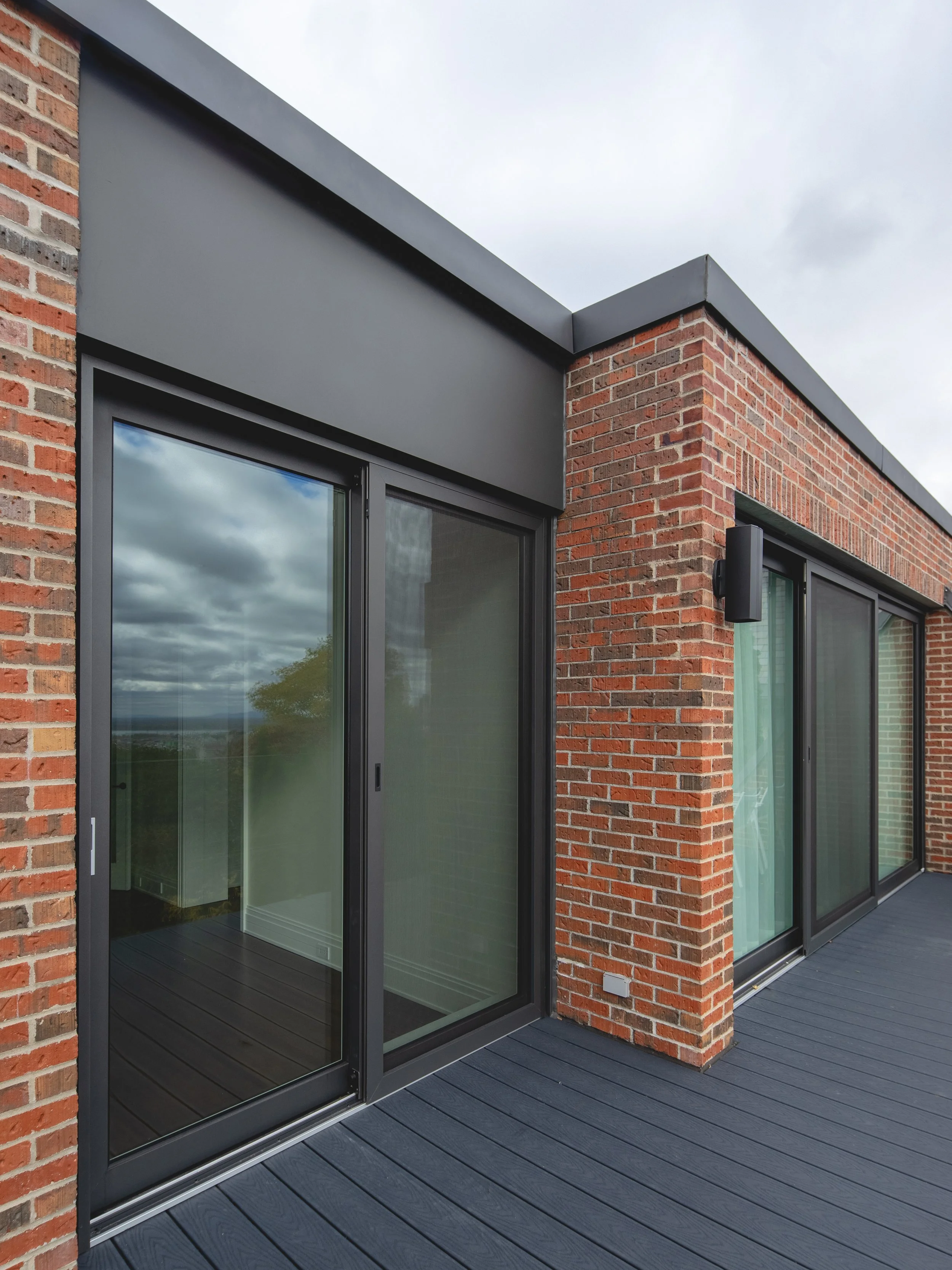 Exterior view of a modern brick building with large sliding glass doors and black metal framing. The balcony has dark gray composite decking, and the sky is overcast.