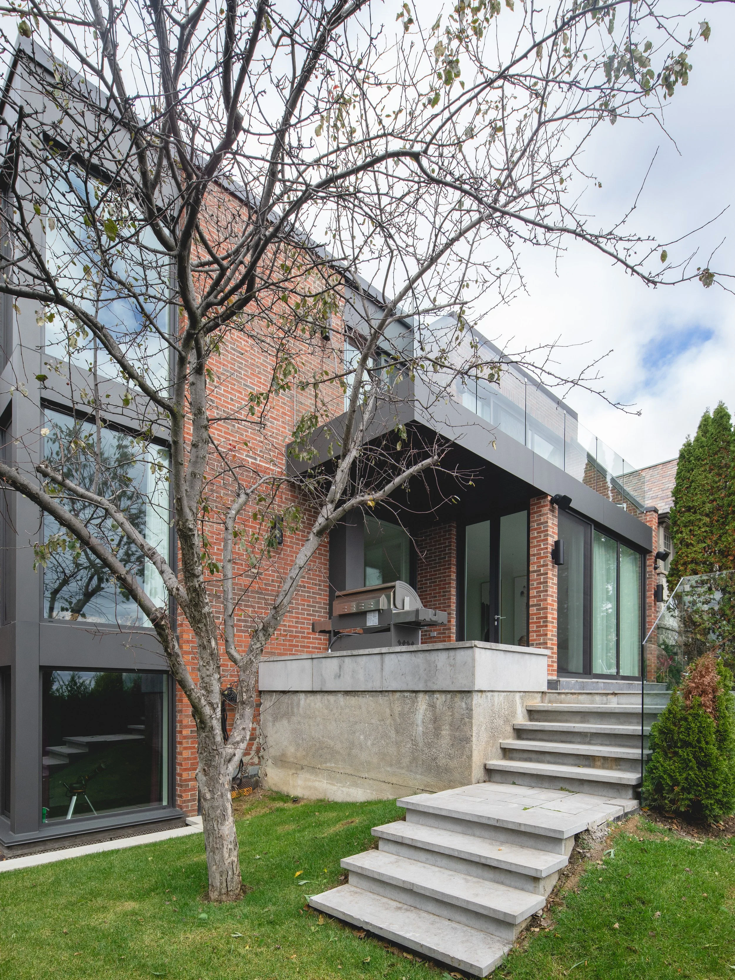 Modern house with brick and black window framing, outdoor concrete stairs leading to a patio with a grill, surrounded by grass and trees with some leaves, under a partly cloudy sky.