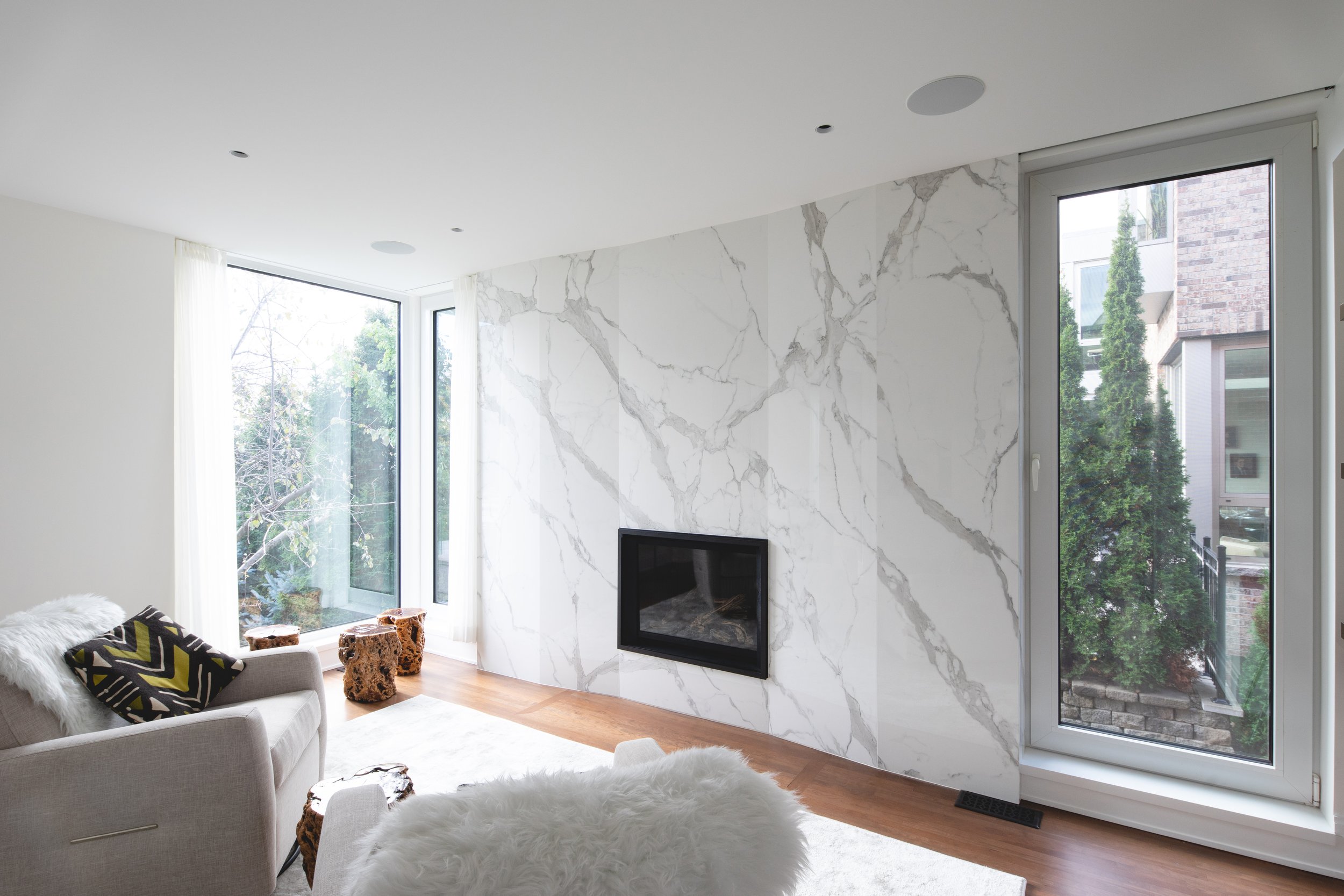 Modern living room with marble accent wall, fireplace, large windows, and minimal furniture including a beige sofa with black and yellow patterned pillows and animal print stools.