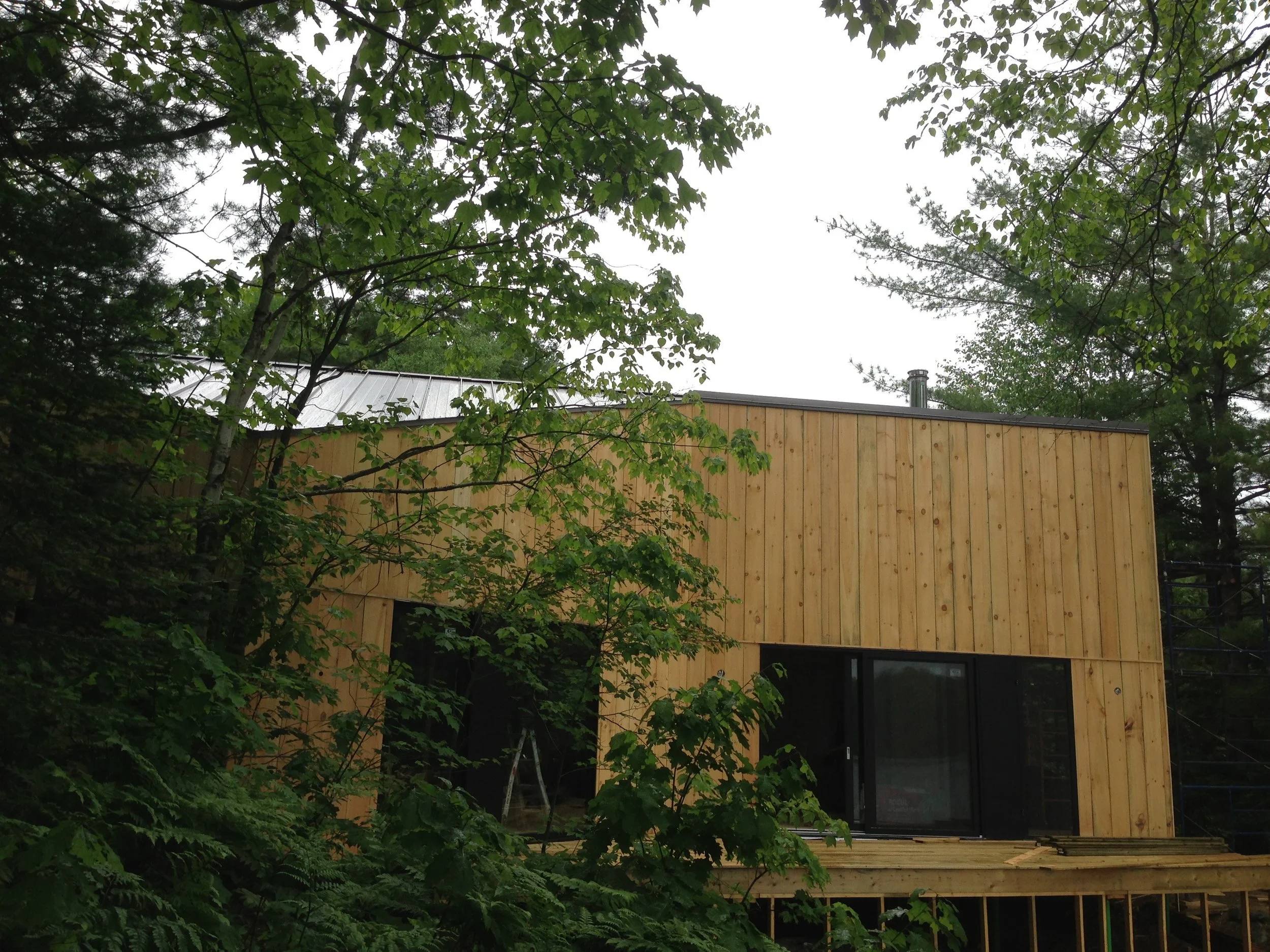 A modern two-story house under construction with wooden exterior siding, surrounded by green trees.