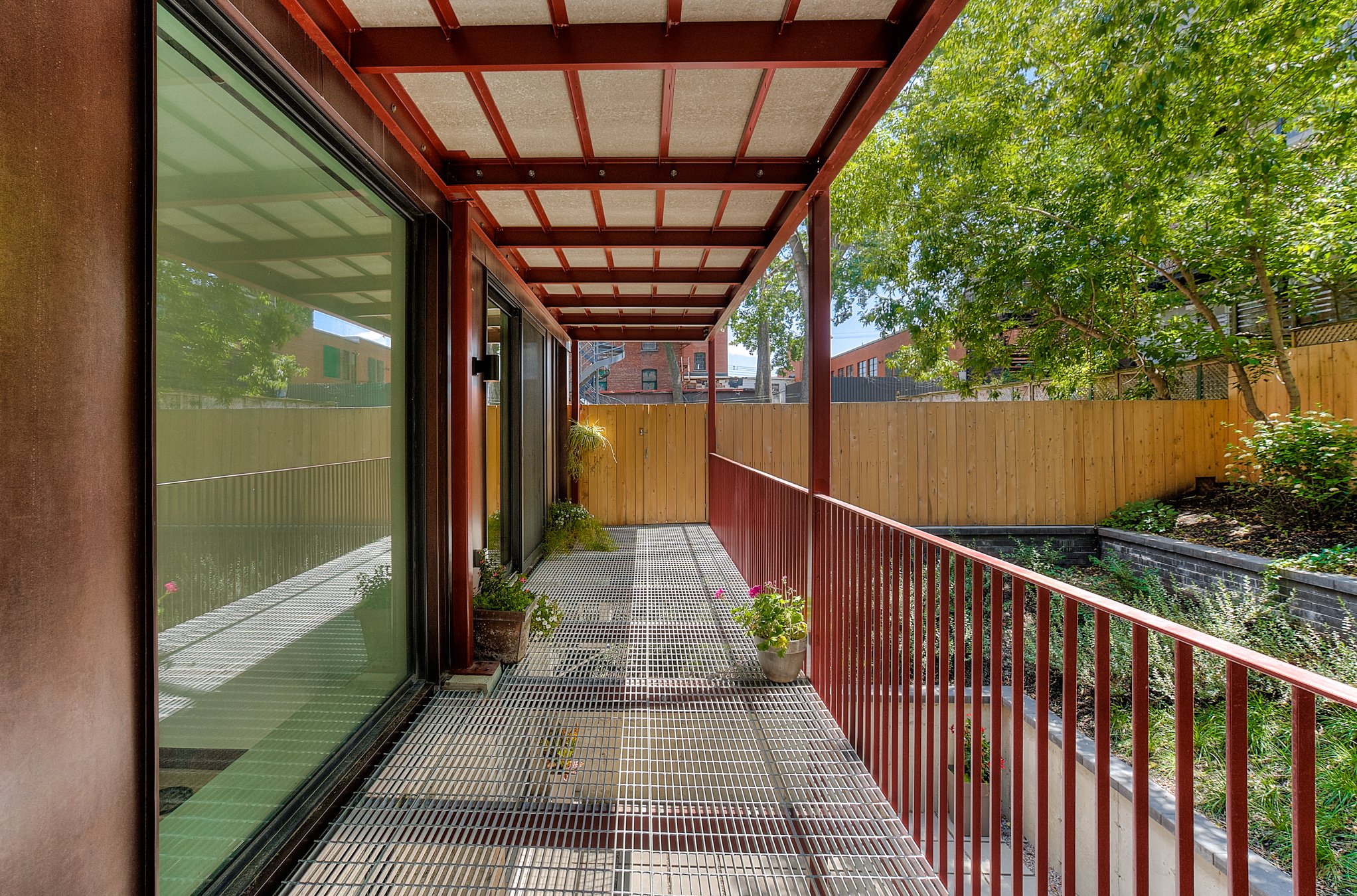 The image shows a narrow balcony with a metal grating floor and a red railing. There are potted plants along the wall, and the balcony is partially shaded by a red metal roof. A wooden fence surrounds the garden area beyond, with trees and greenery v