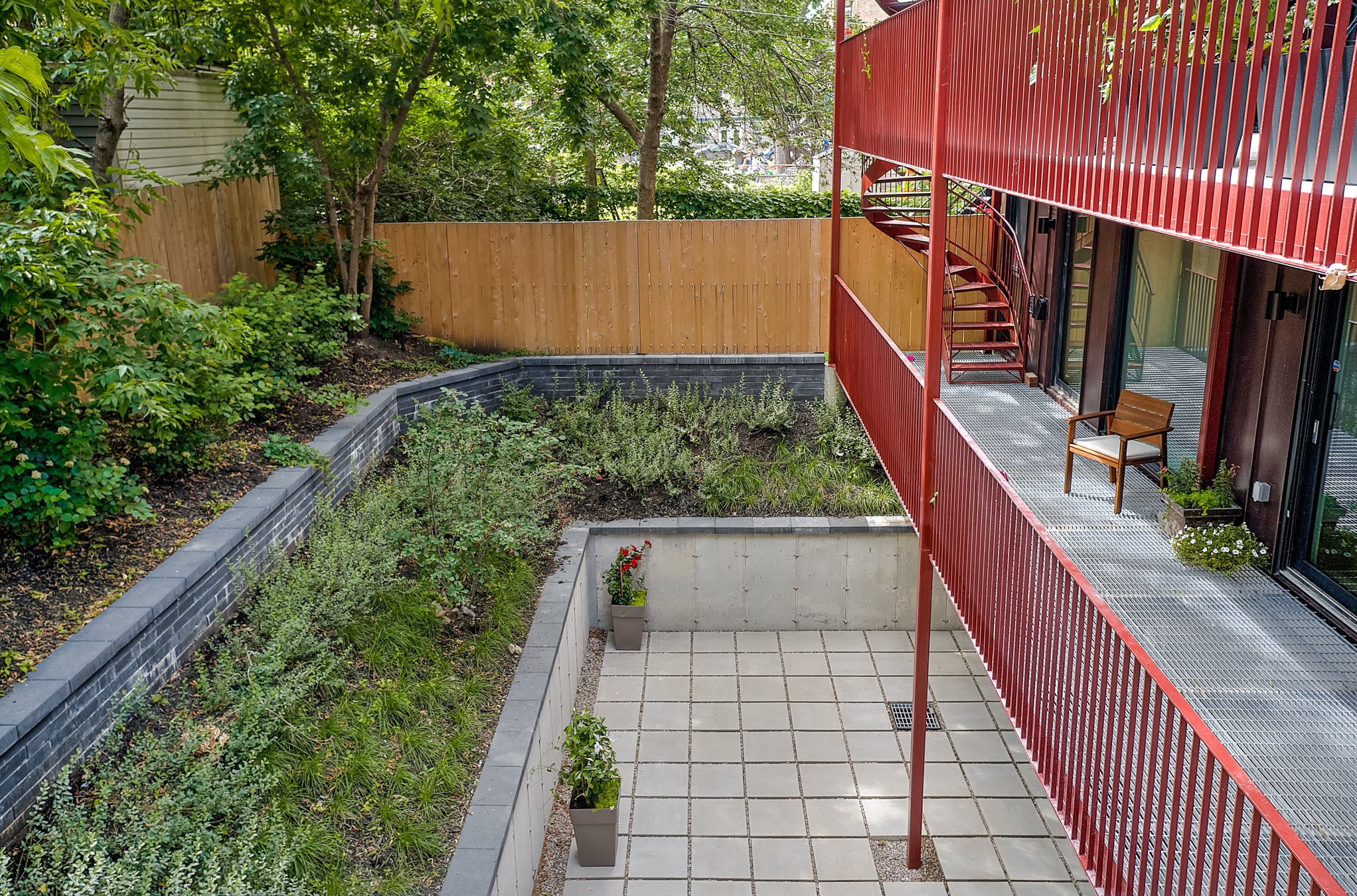 A modern outdoor balcony with red railing, a wooden chair, potted plants, and a stairway leading to the upper level. The yard has a tiled patio, a wooden fence, and lush green trees and shrubs in the background.