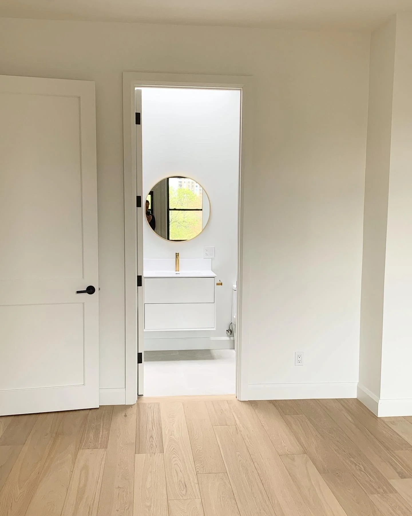 View through a doorway into a modern bathroom with a white vanity, a round mirror, and a window showing trees outside.