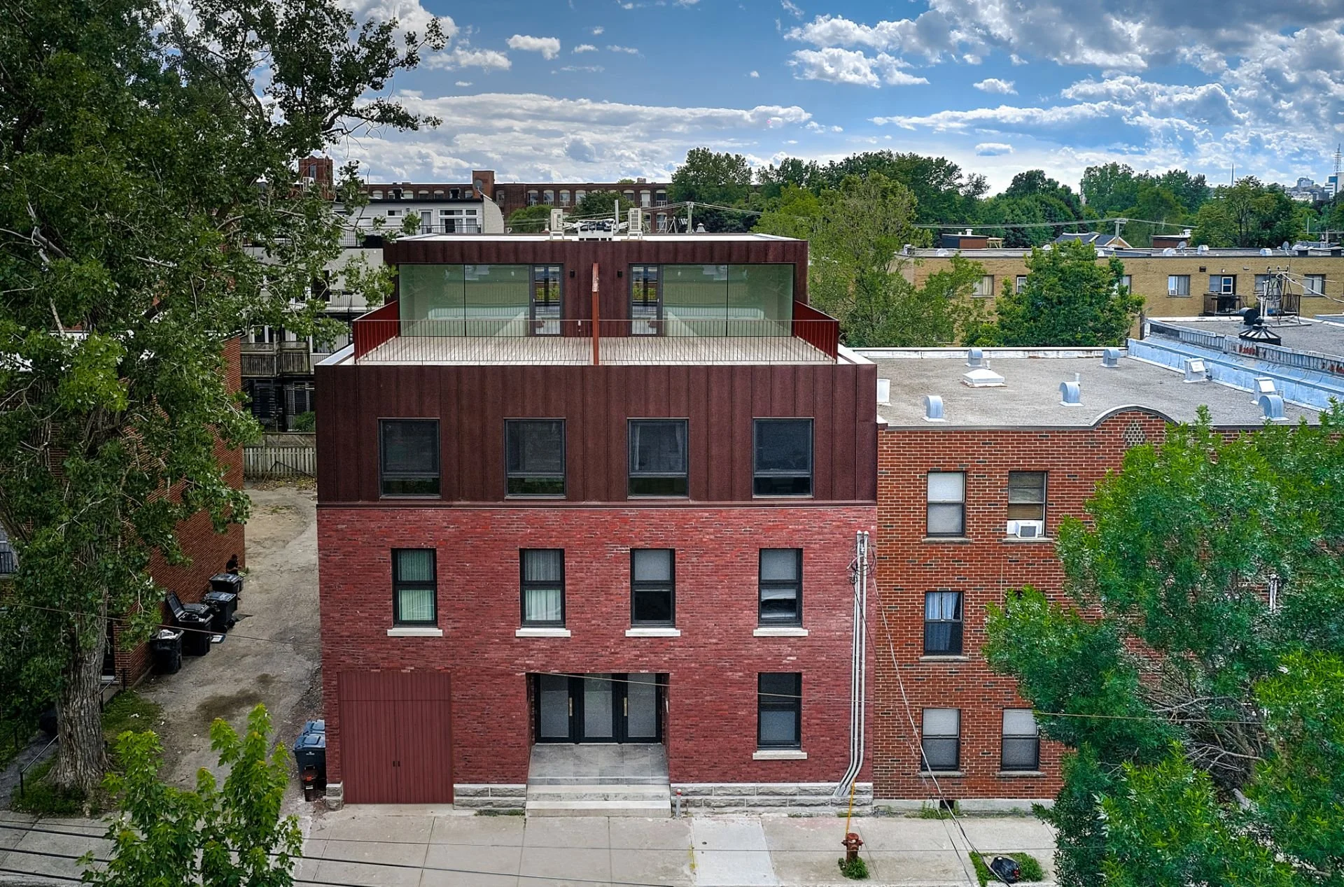 A multi-story brick building with a rooftop terrace, large glass windows, and a red metal facade on the upper level, surrounded by trees and neighboring buildings under a partly cloudy sky.