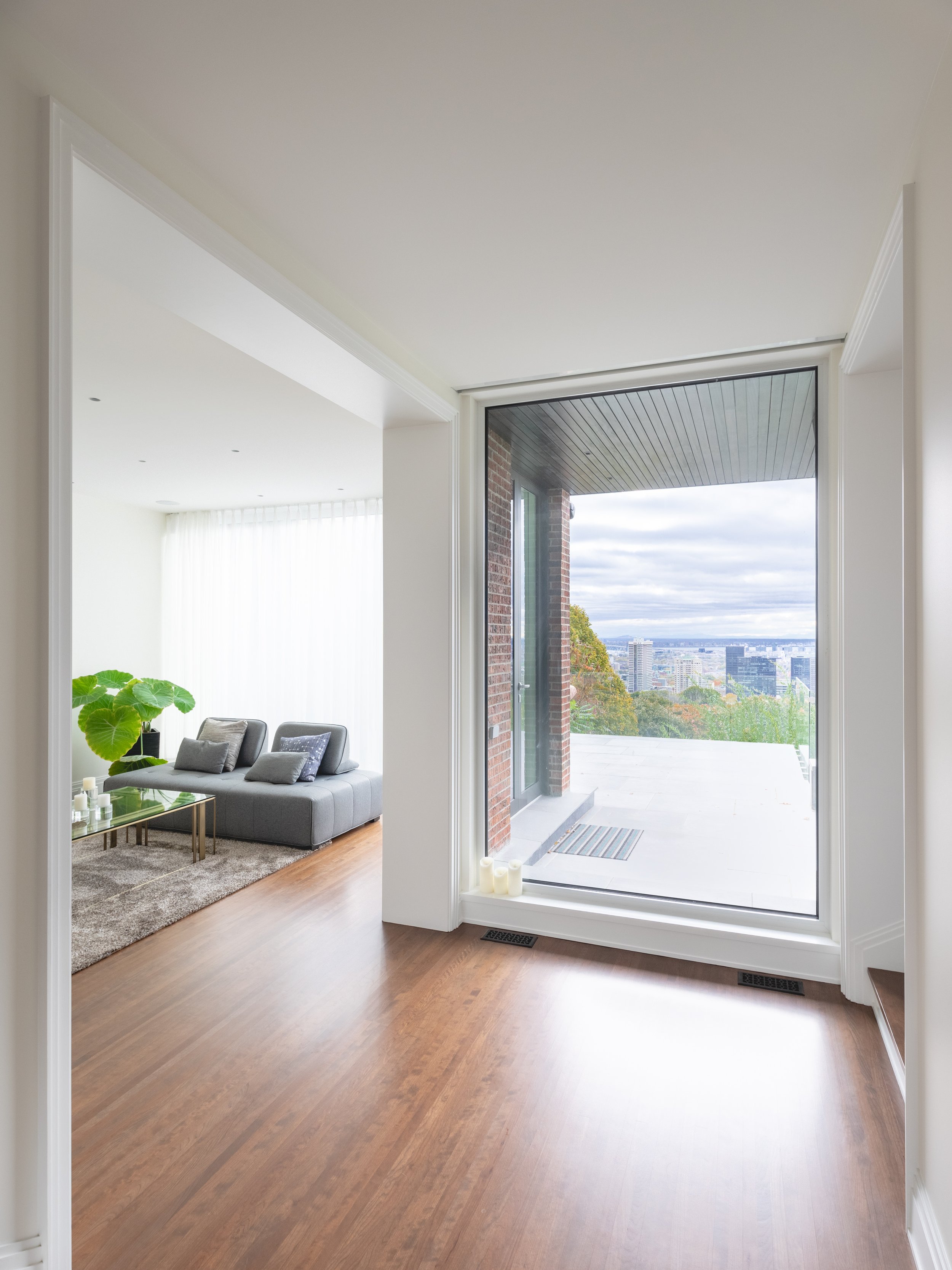 Interior view of a modern apartment with wooden flooring, a large sliding glass door leading to a balcony, and a living room with a gray sofa, a glass coffee table, and large potted plant.