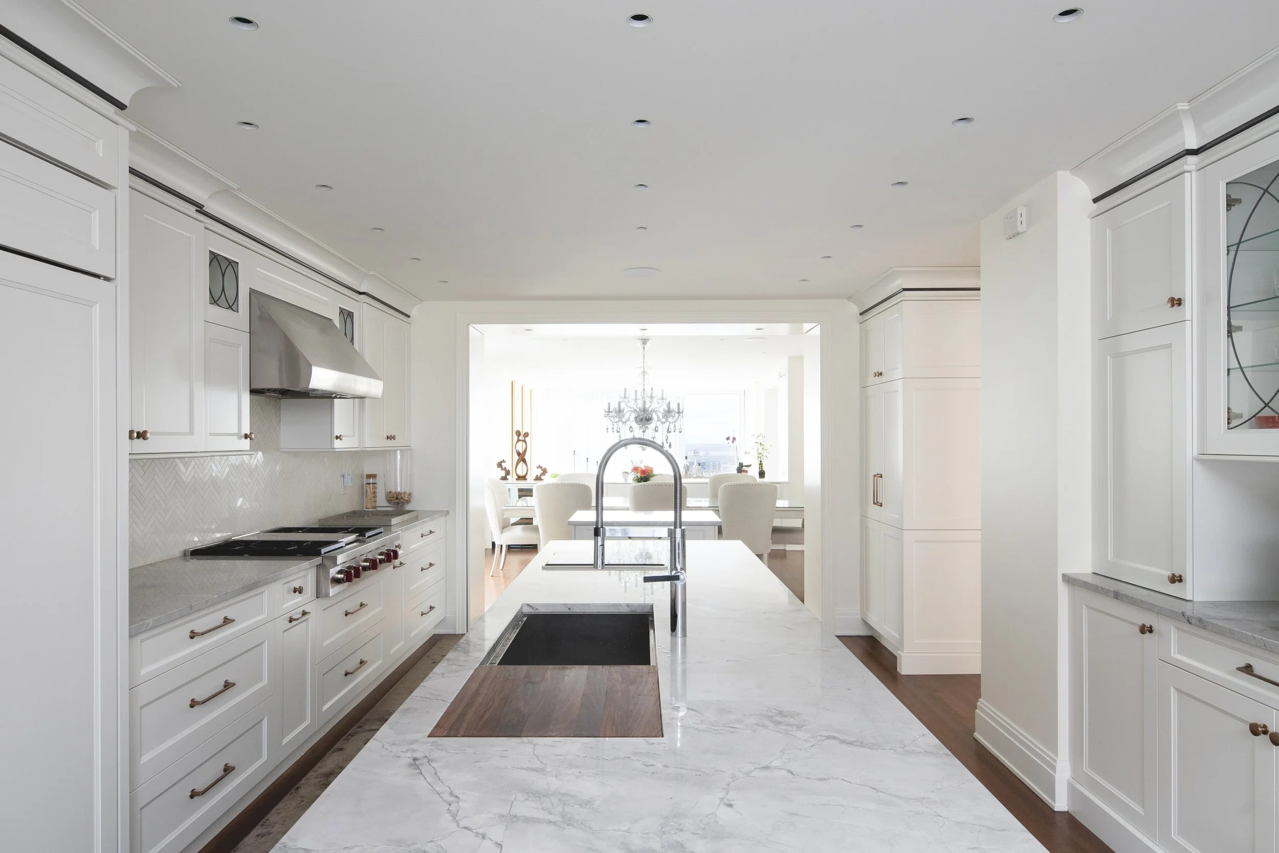 Modern, white kitchen with marble countertop island, stainless steel stove, and a view of a bright dining area with chandelier in the background.