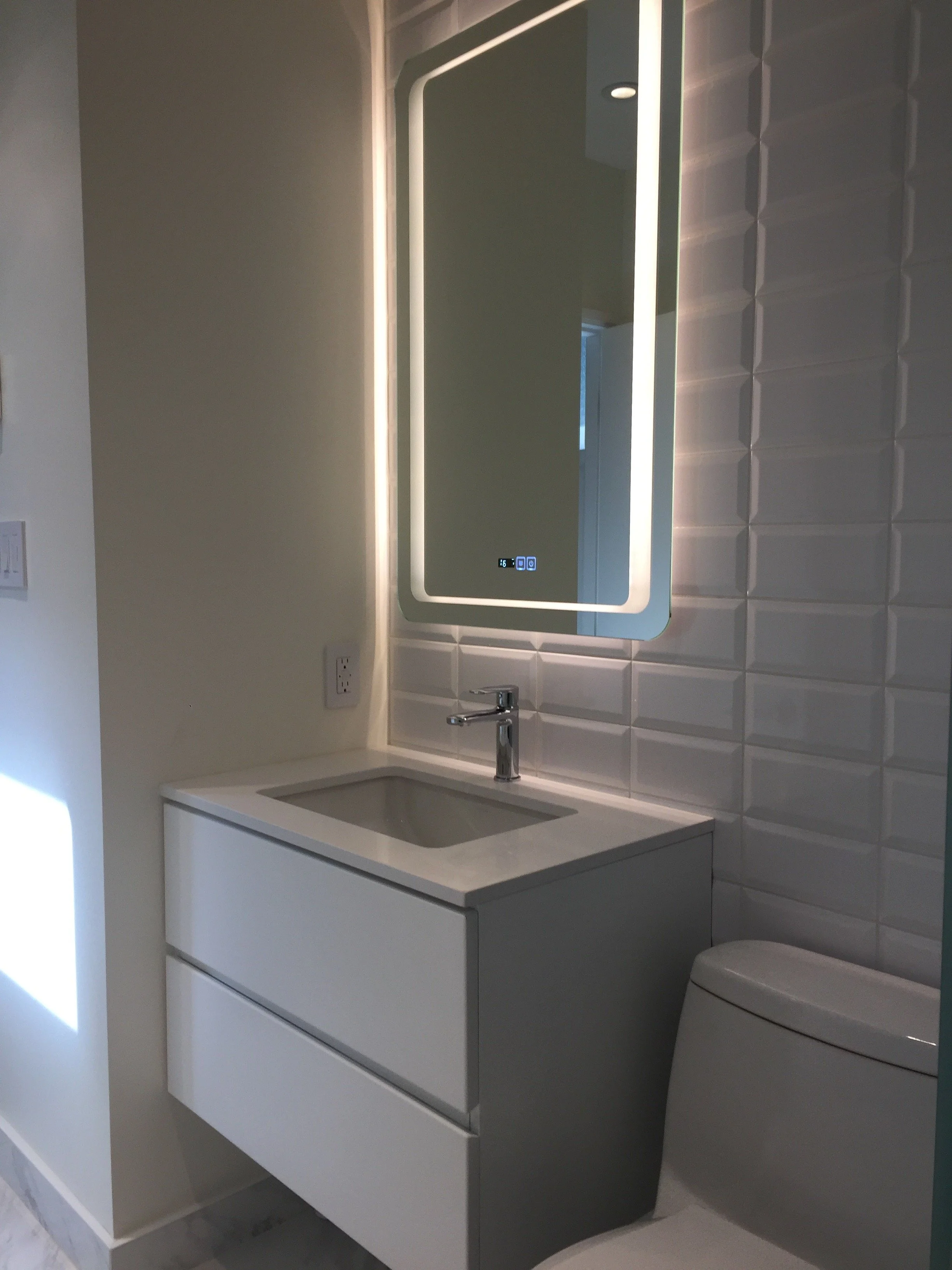 Modern bathroom with a wall-mounted white sink cabinet, a rectangular mirror with LED lighting, a sleek faucet, and a white toilet. The wall behind the sink features textured white tiles.