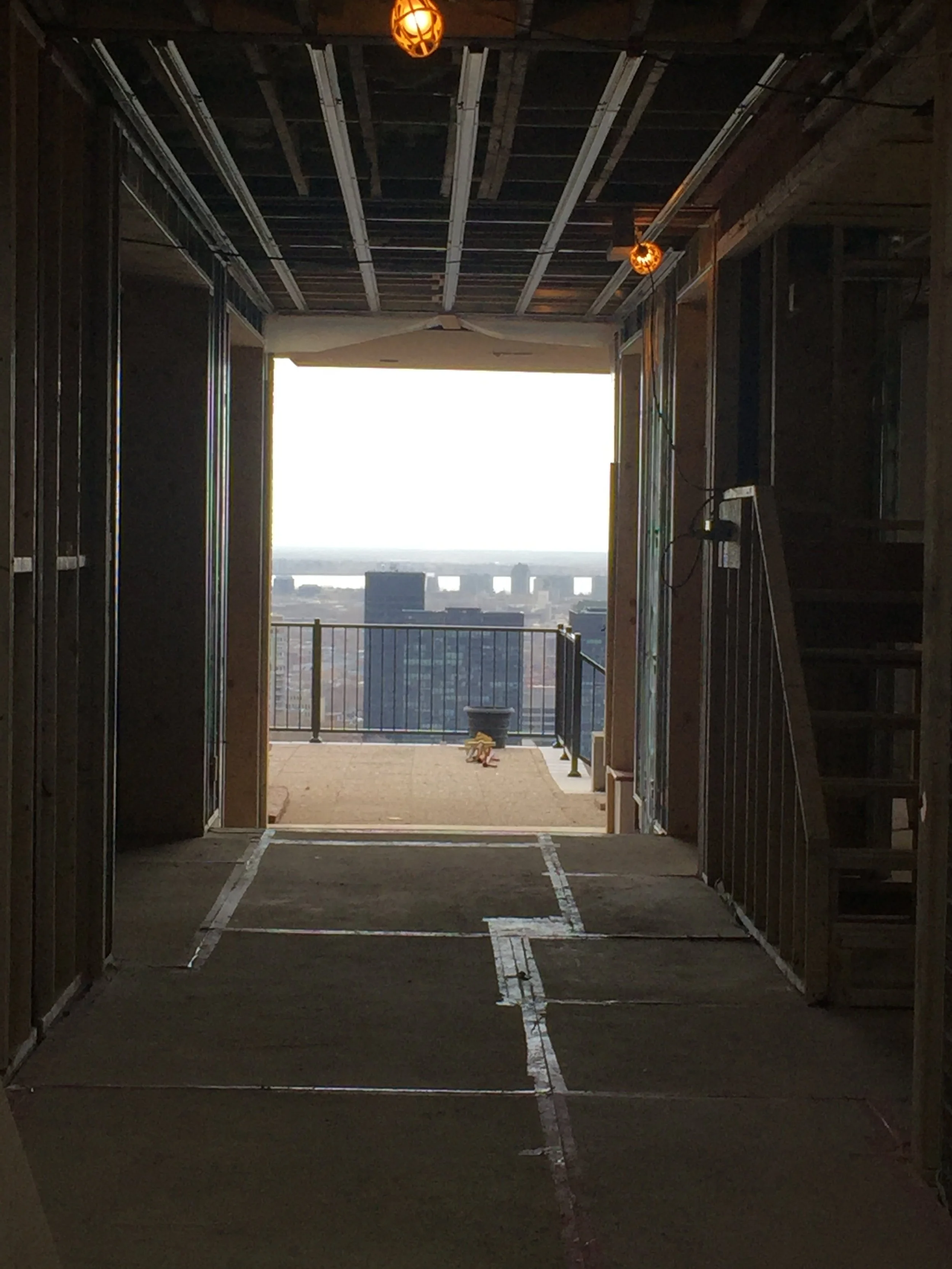 View from a high-rise balcony under construction, showing an unfinished interior with exposed walls and ceiling, and a city skyline in the distance through a large opening.
