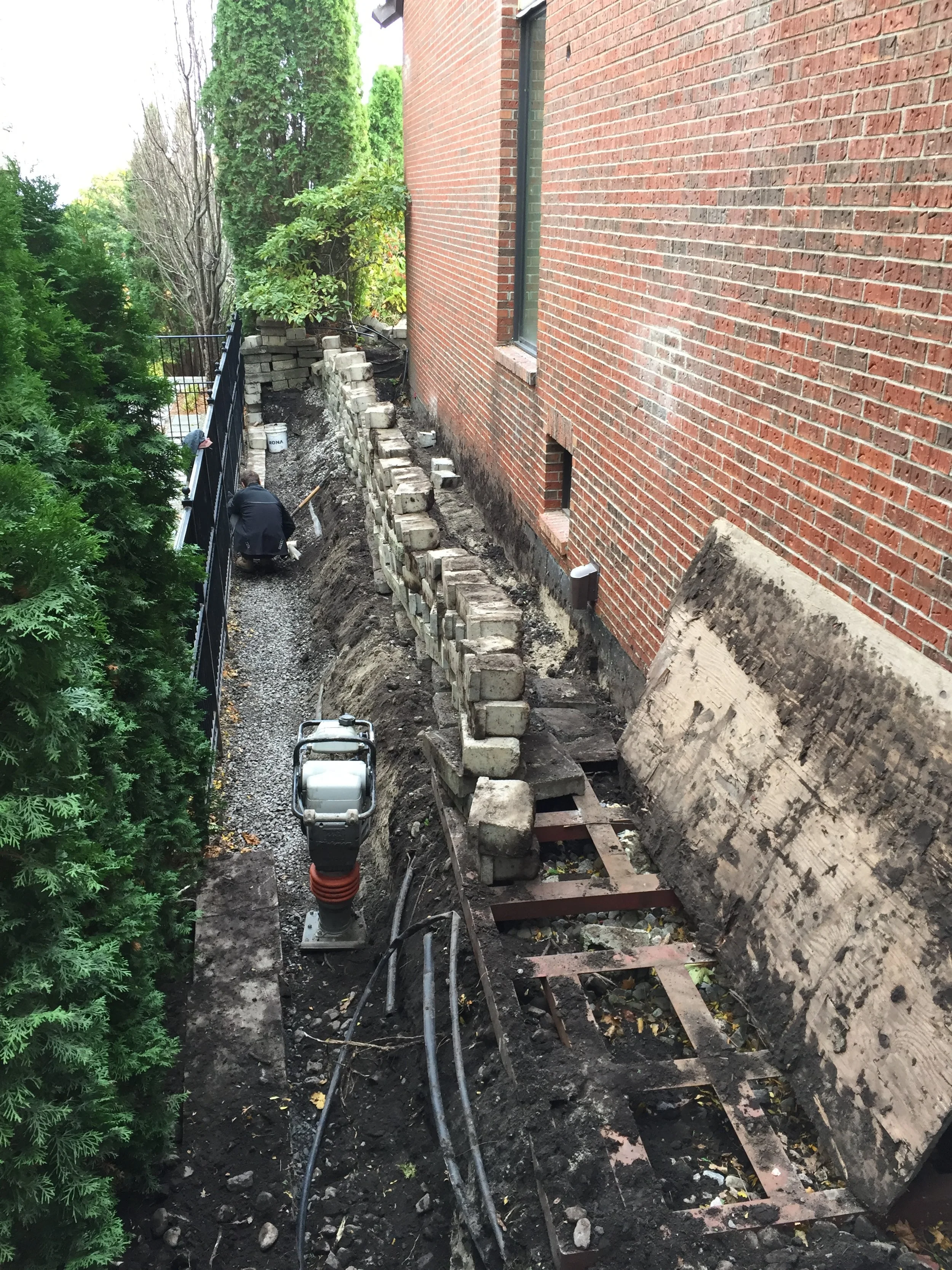 A construction worker is building a retaining wall with bricks along the side of a brick building. Construction equipment and scattered bricks are present in the area.