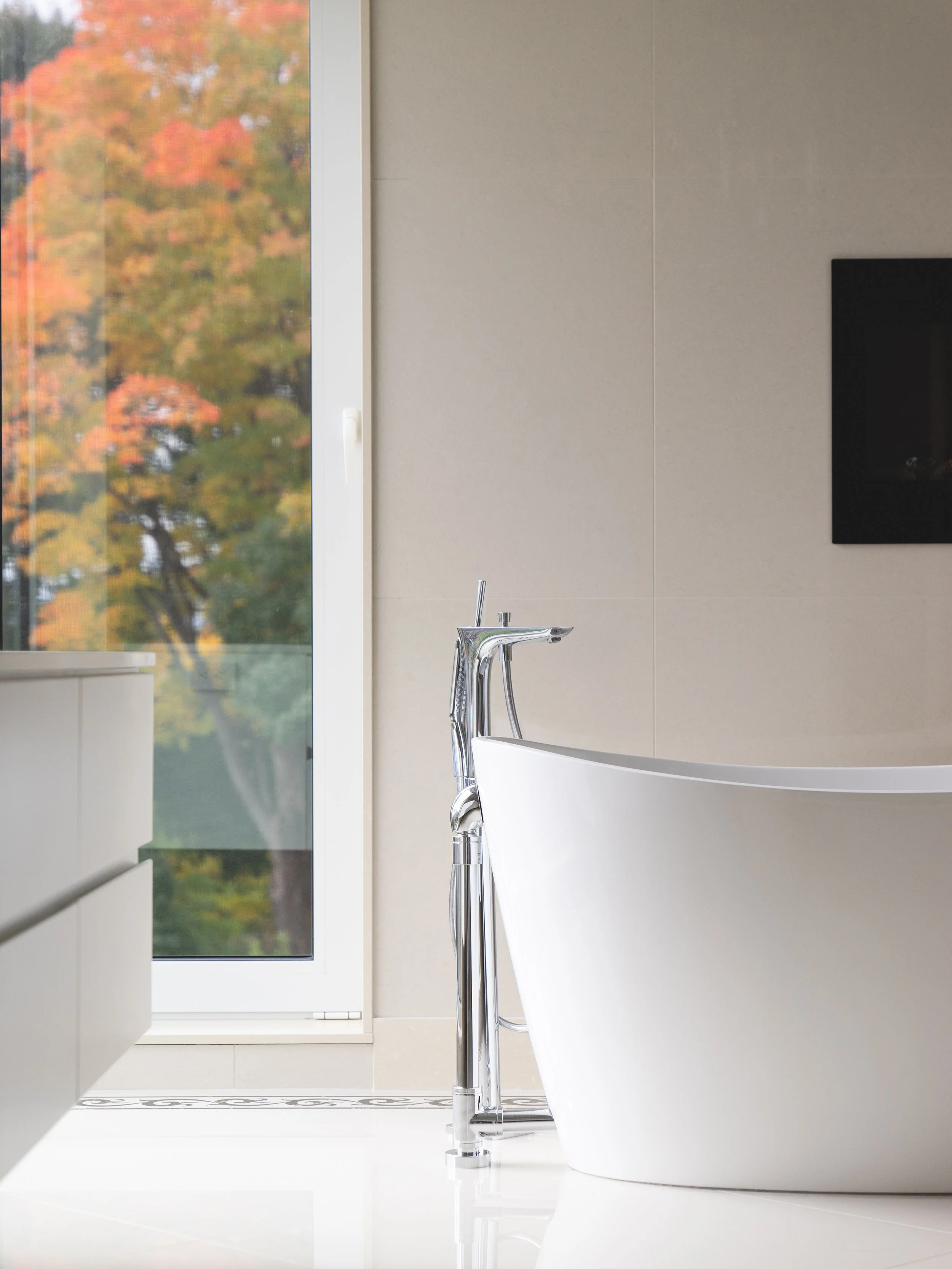 Modern white freestanding bathtub with a chrome floor-mounted faucet in a bright bathroom, with open window showing autumn trees.