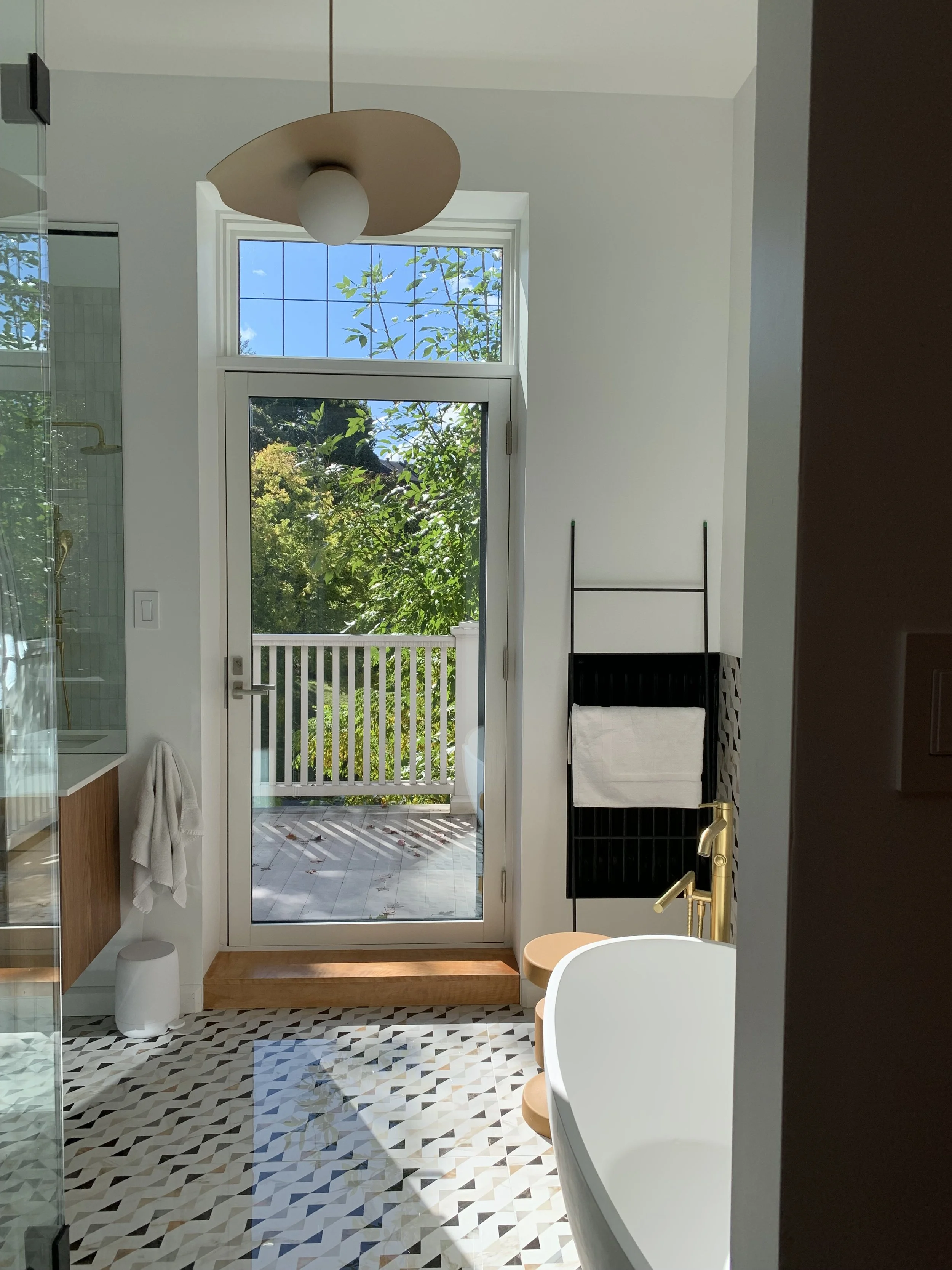 Bathroom with a glass door leading to a balcony, a freestanding bathtub, a black towel rack with a white towel, a wooden step, and patterned tile floor, with outdoor plants visible through the window.